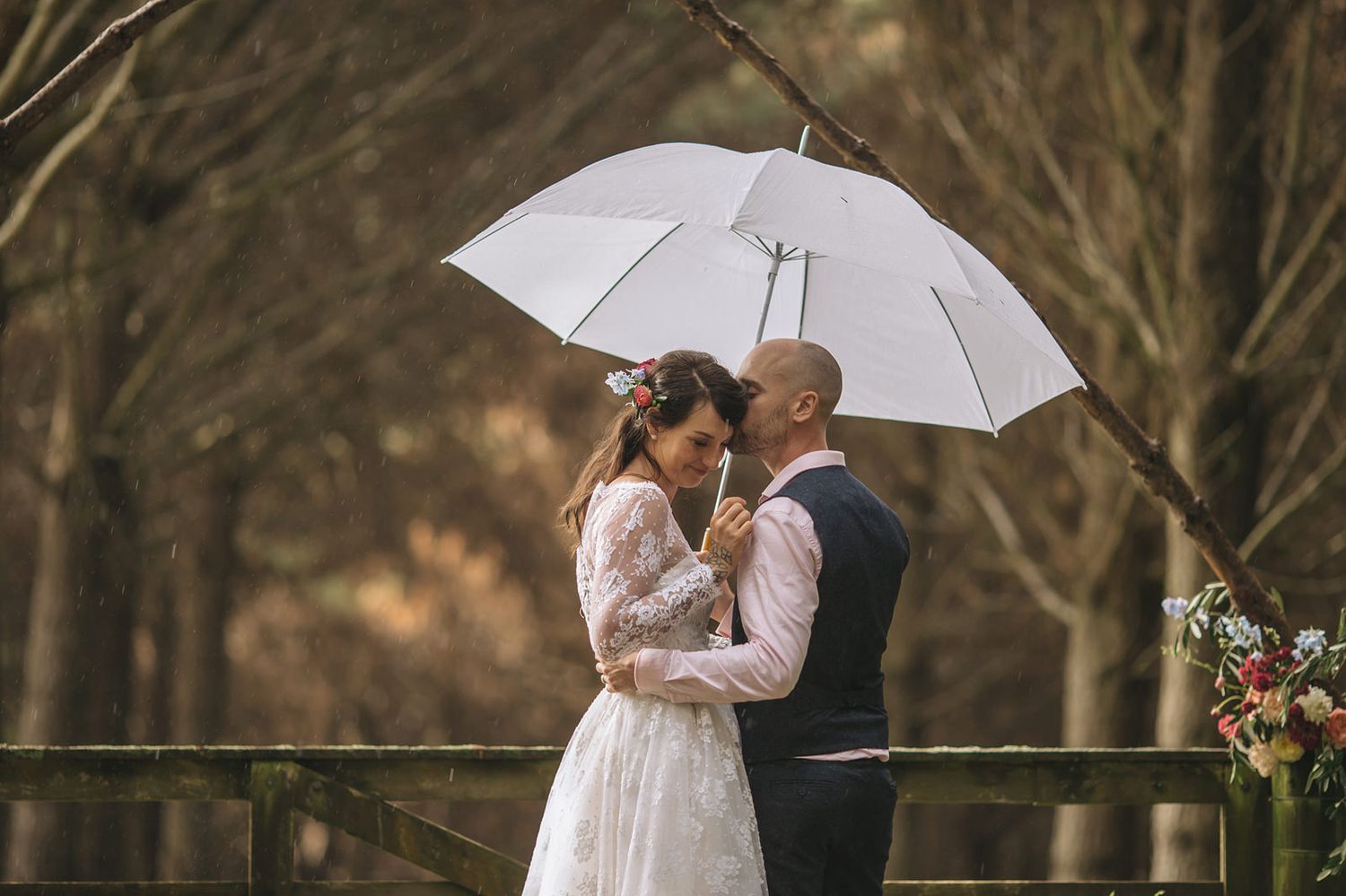 Couple shares romantic embrace under white umbrella during outdoor wedding photoshoot in wooded setting.