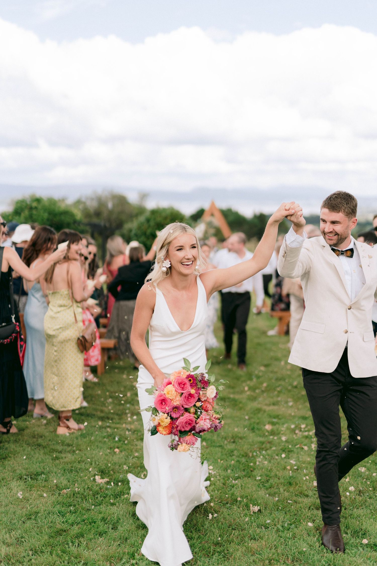 Newlyweds celebrate with joyful dance on grassy field with guests in background.