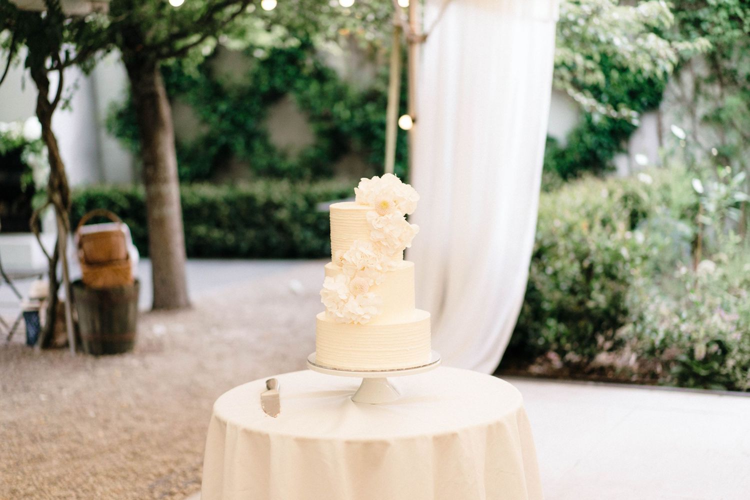 Elegant three-tier white wedding cake displayed on a round table with flowing fabric backdrop.