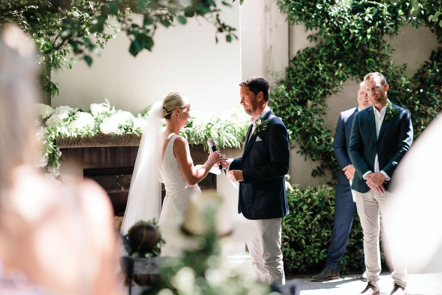 Outdoor wedding ceremony with floral decor and greenery against white walls.