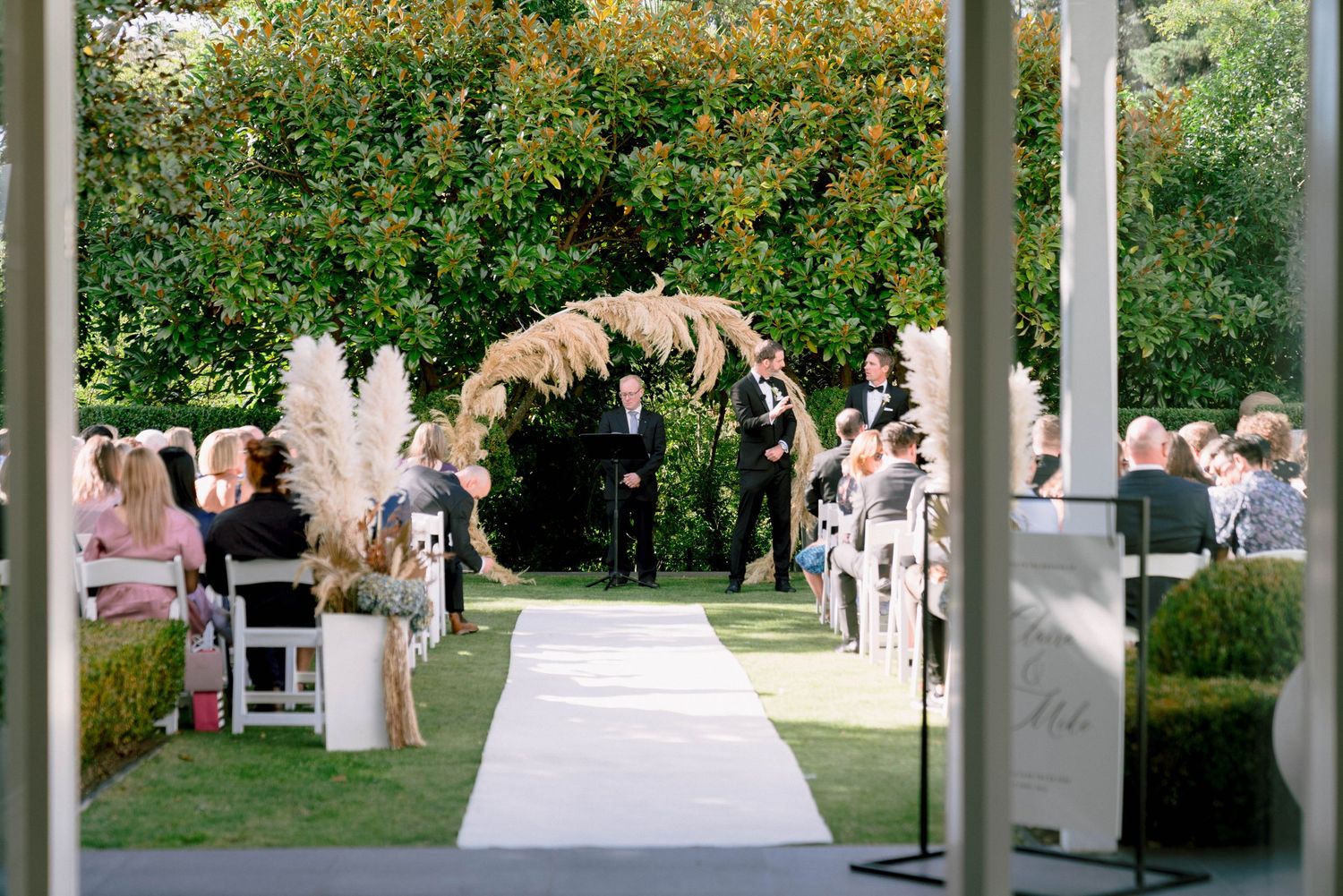 Outdoor wedding ceremony setup with a floral arch over white aisle runner and guests seated on white chairs in garden setting.
