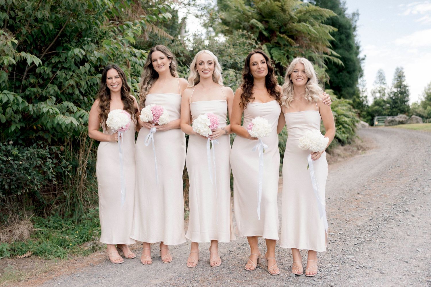 Five bridesmaids in matching cream colored dresses pose together on country road.