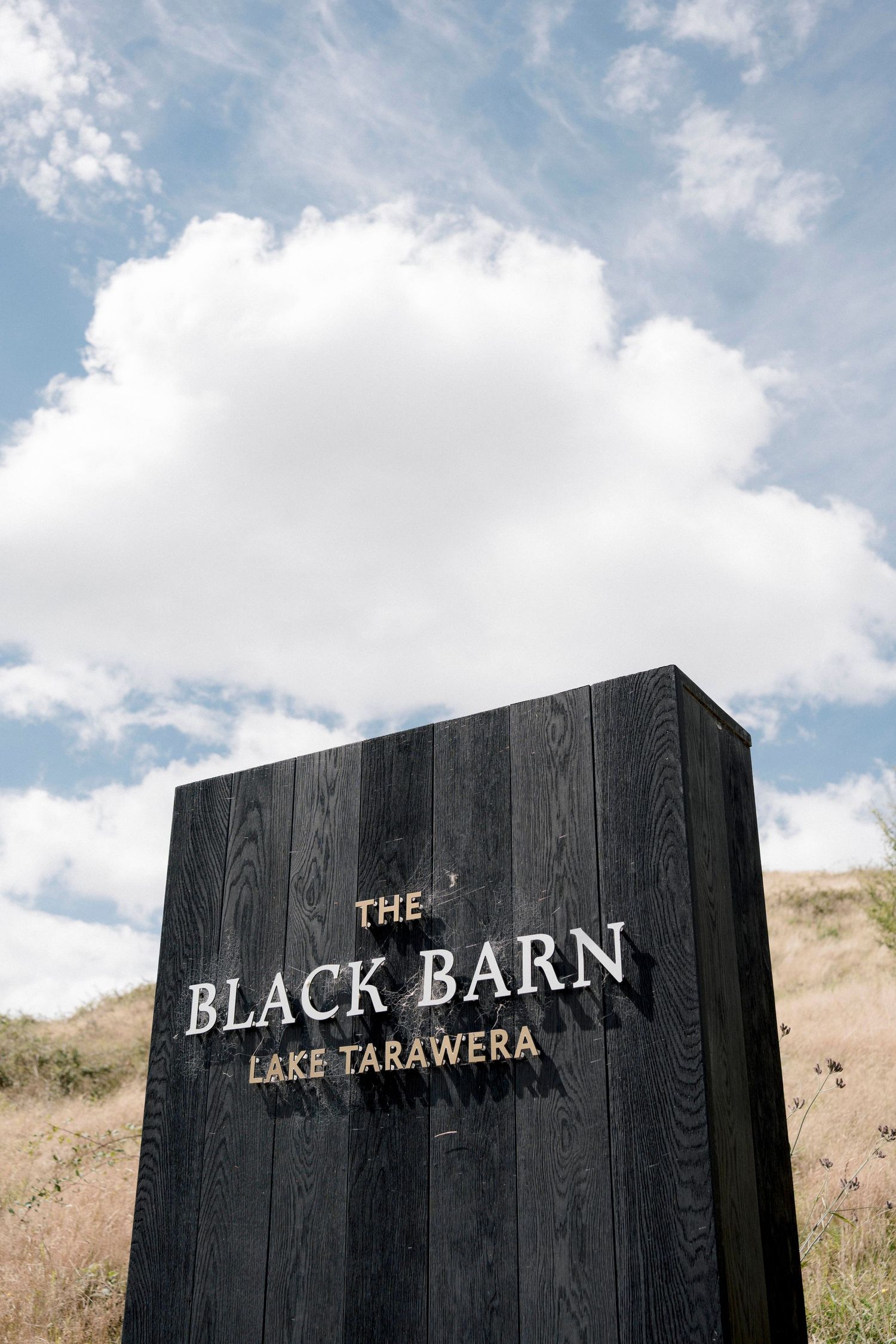 A black wooden sign for 'The Black Barn Lake Tarawera' against a cloudy blue sky.
