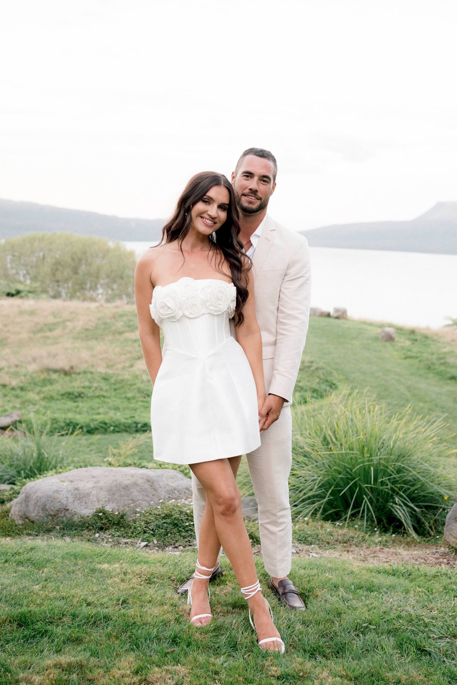 A couple in matching white outfits poses together on a scenic hillside overlooking a serene lake.