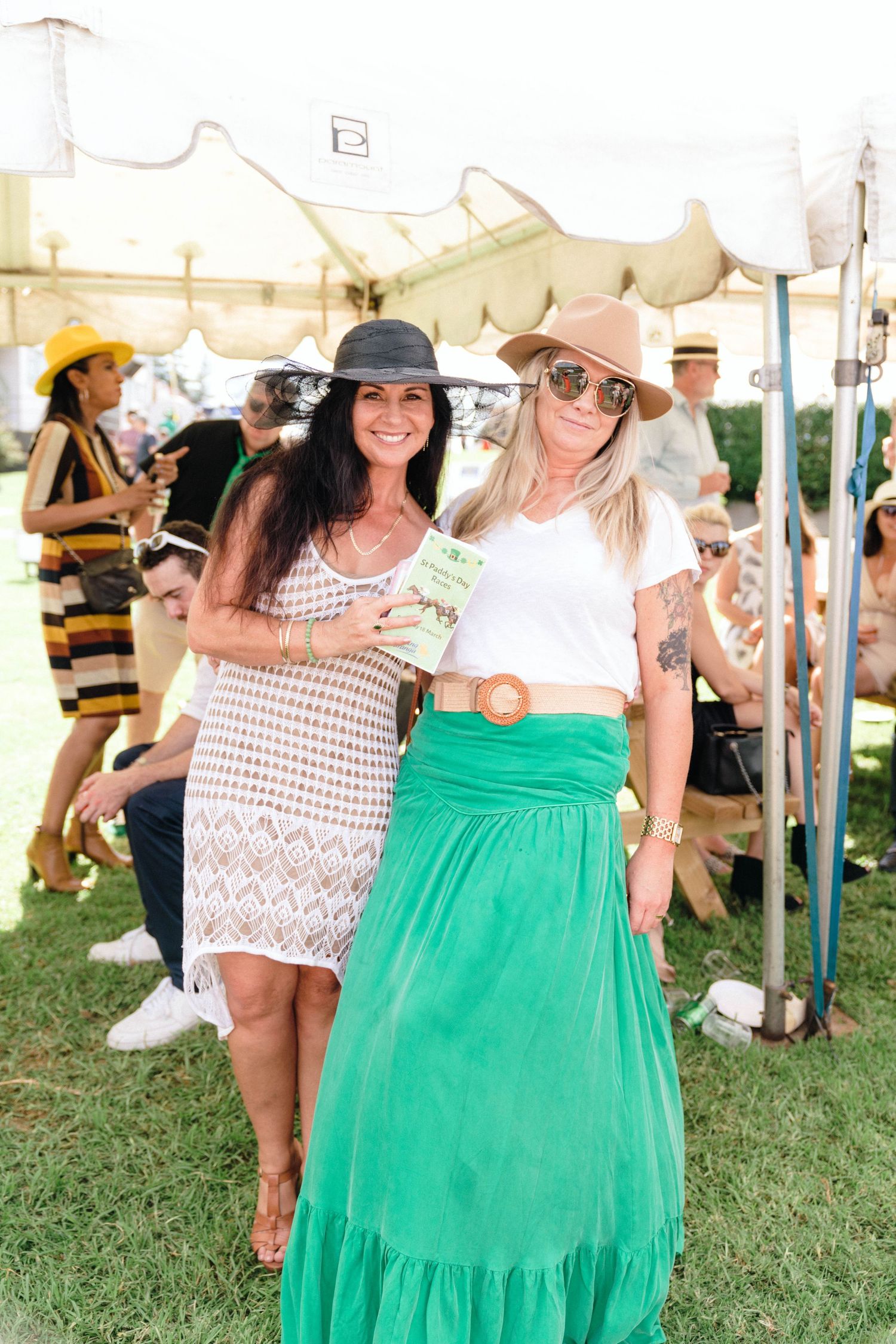 Friends in sundresses and summer hats enjoy a sunny day at an outdoor gathering with a green tent visible.