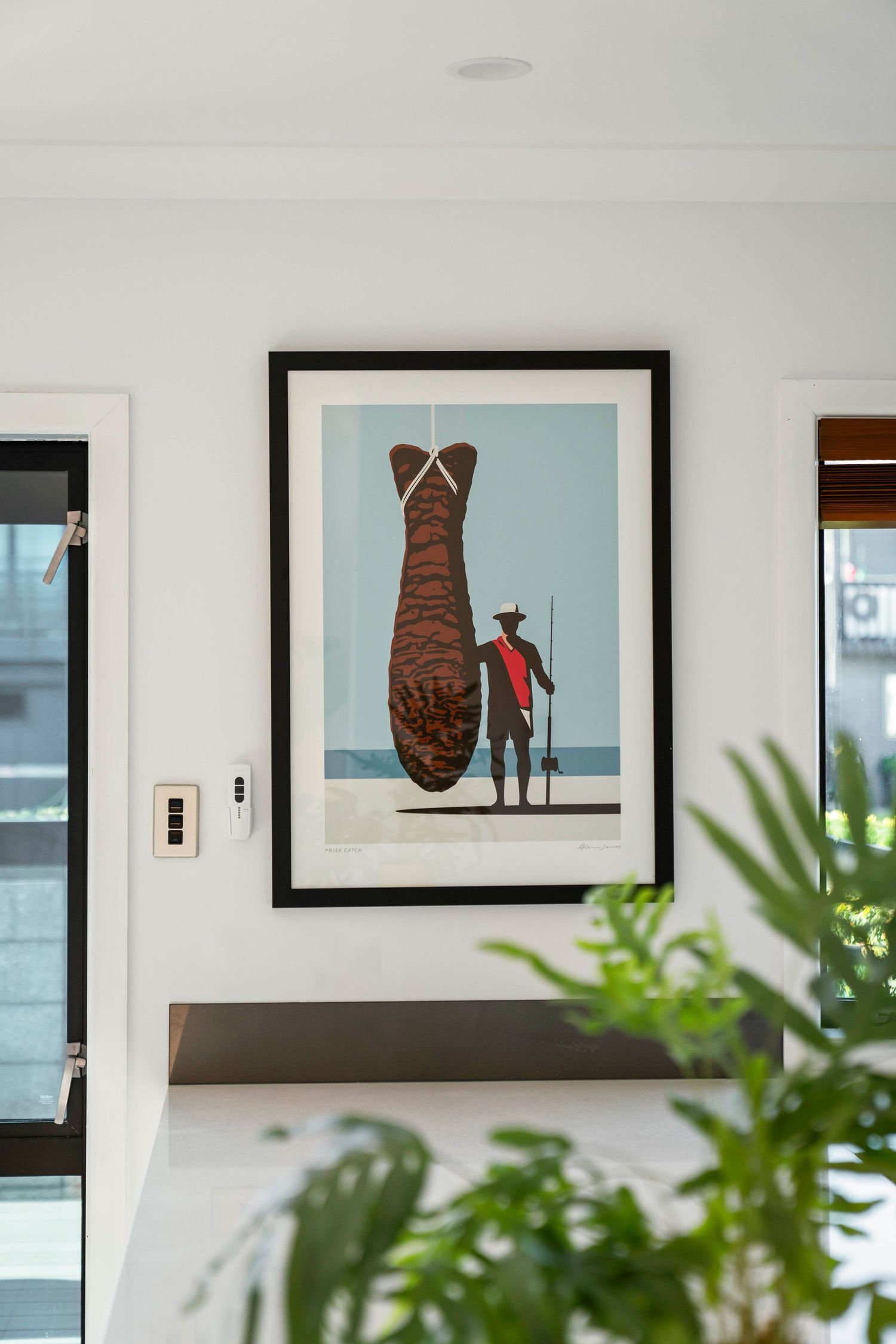 Framed artwork featuring whimsical illustration of large brown fish with small figure on white wall.