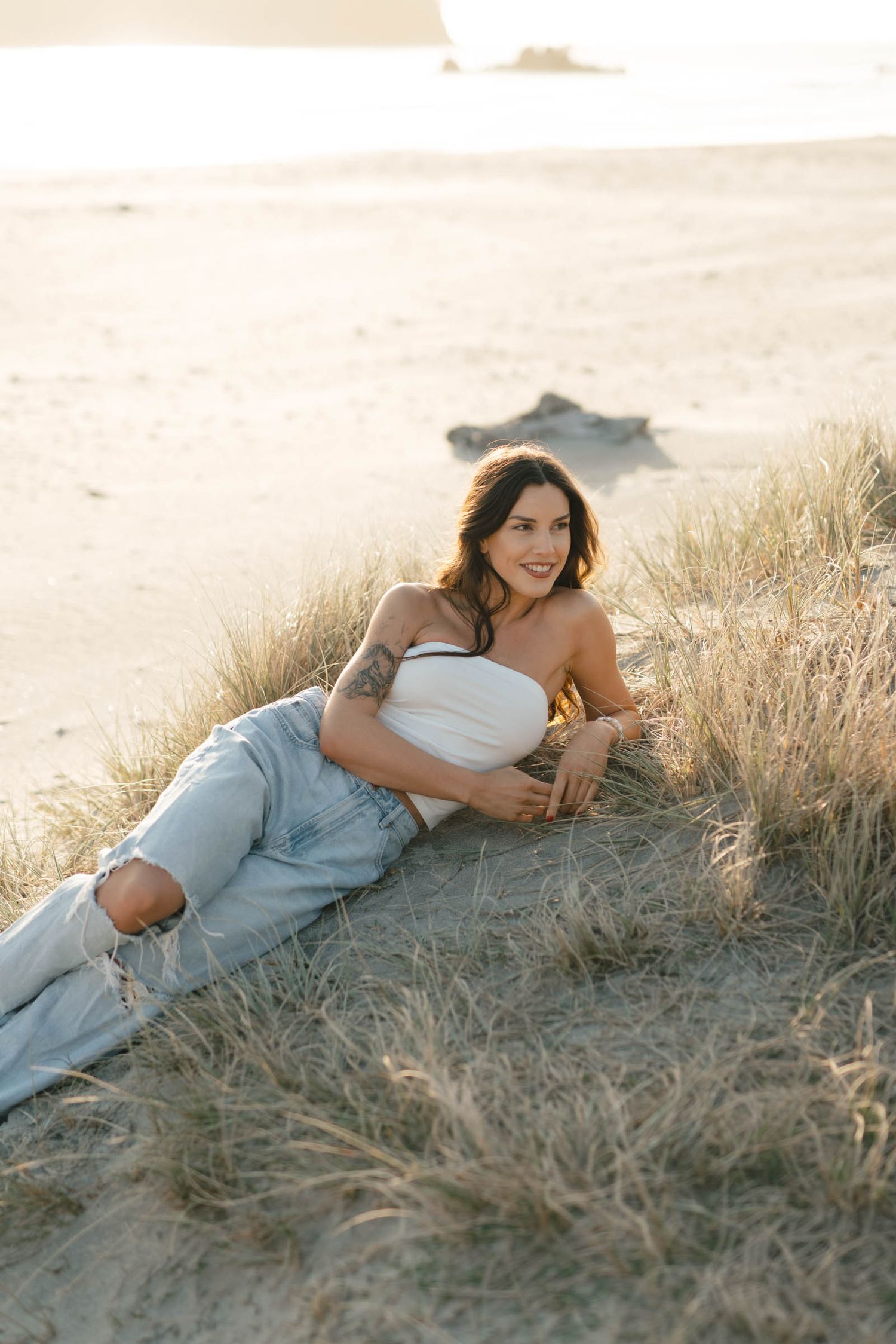 Person in light clothing reclines in golden grass during a dreamy beach photoshoot.
