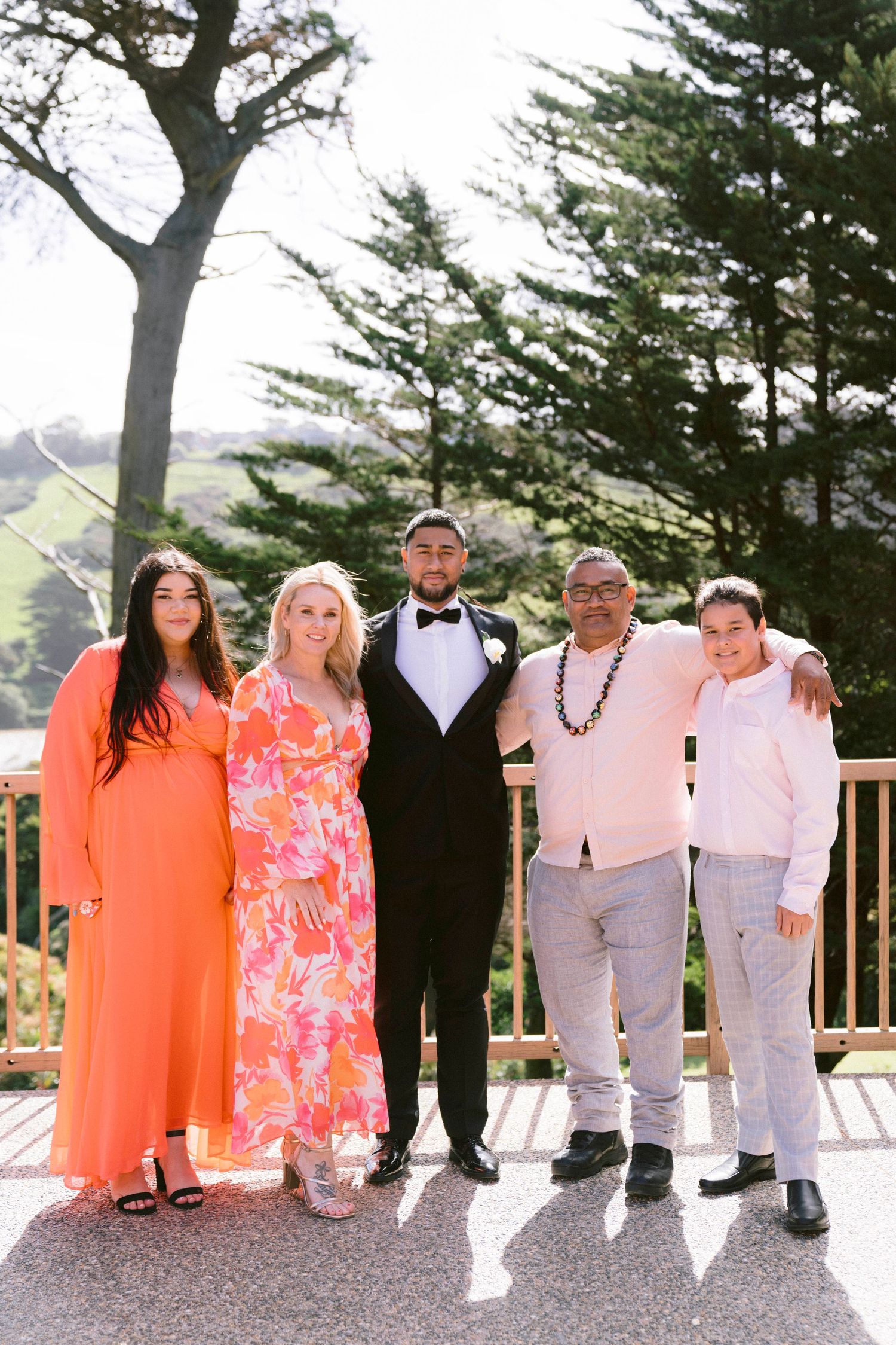Wedding guests in bright coral and white outfits pose together on outdoor deck.