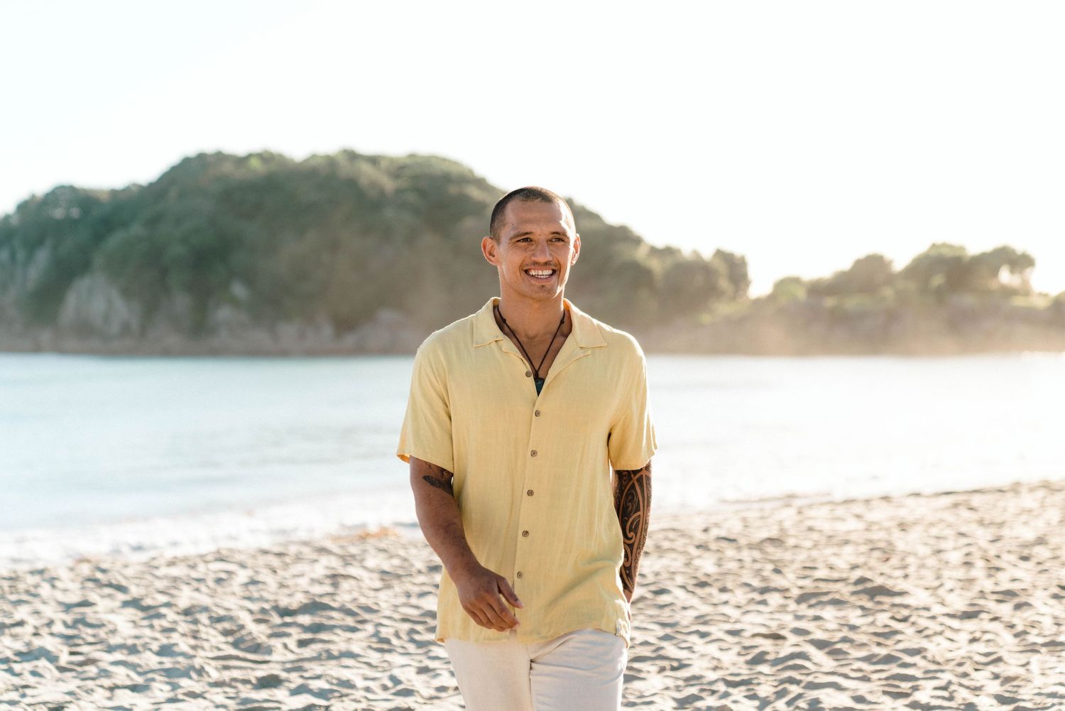 Person in yellow button-down shirt and white pants smiling while standing on sunny beach with coastal cliffs behind.