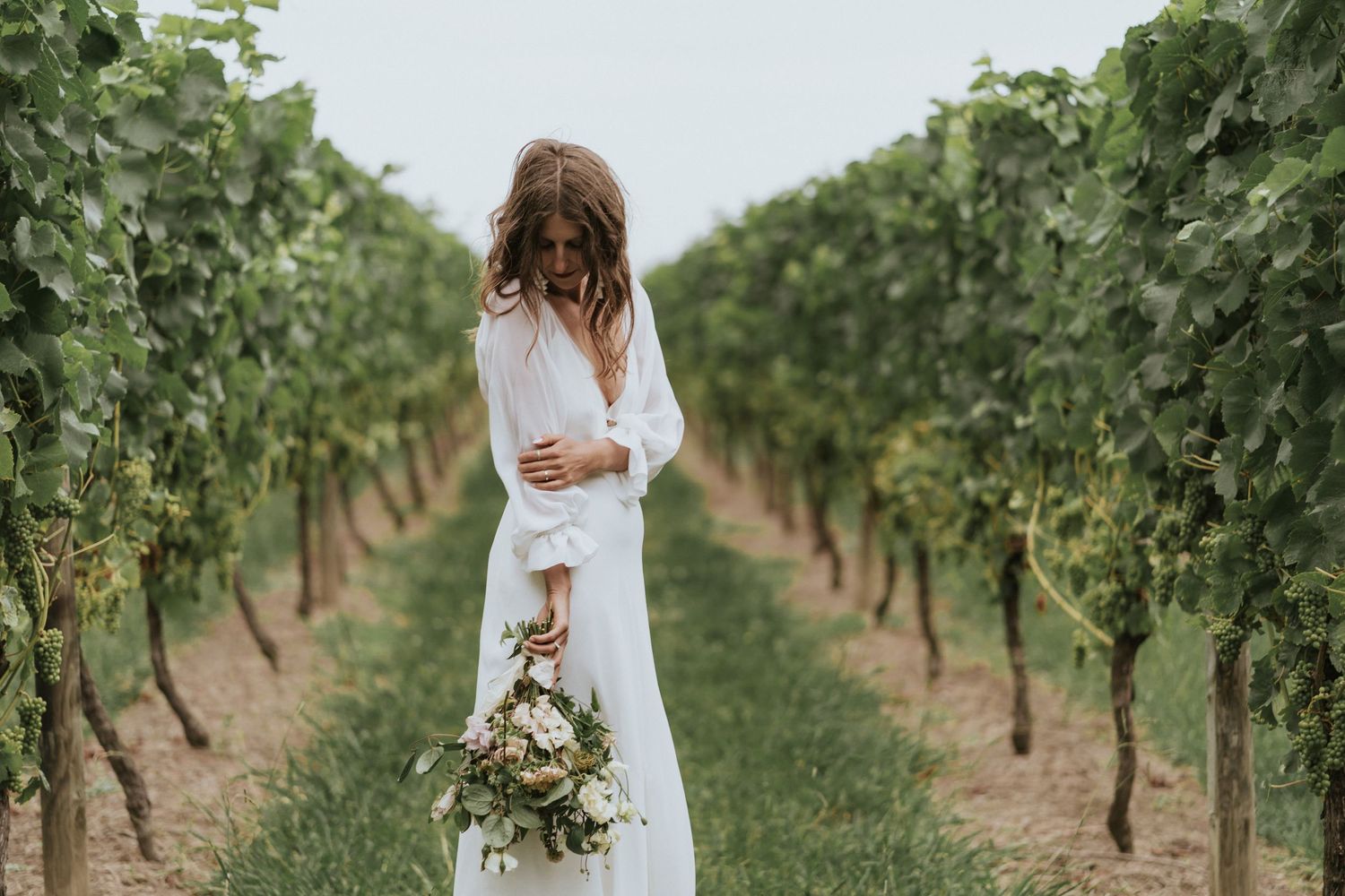 A person in a white dress stands in a vineyard holding a bouquet while surrounded by rows of grapevines.