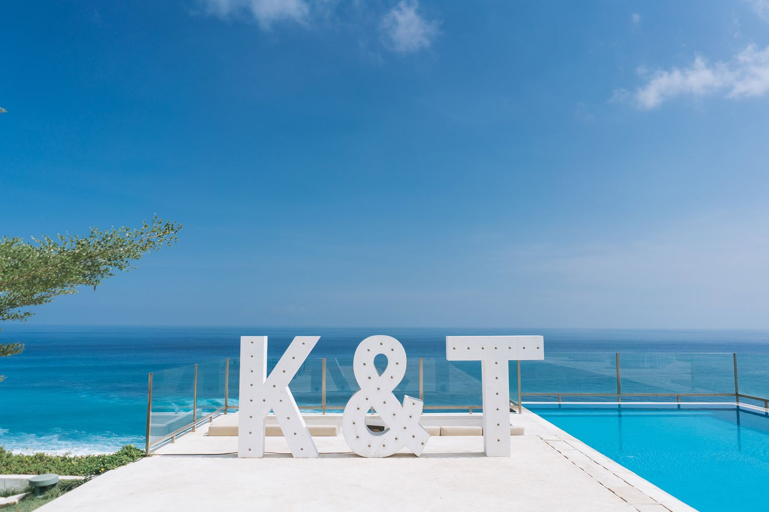 Large white K&T letters stand against infinity pool overlooking tropical ocean under bright blue sky.
