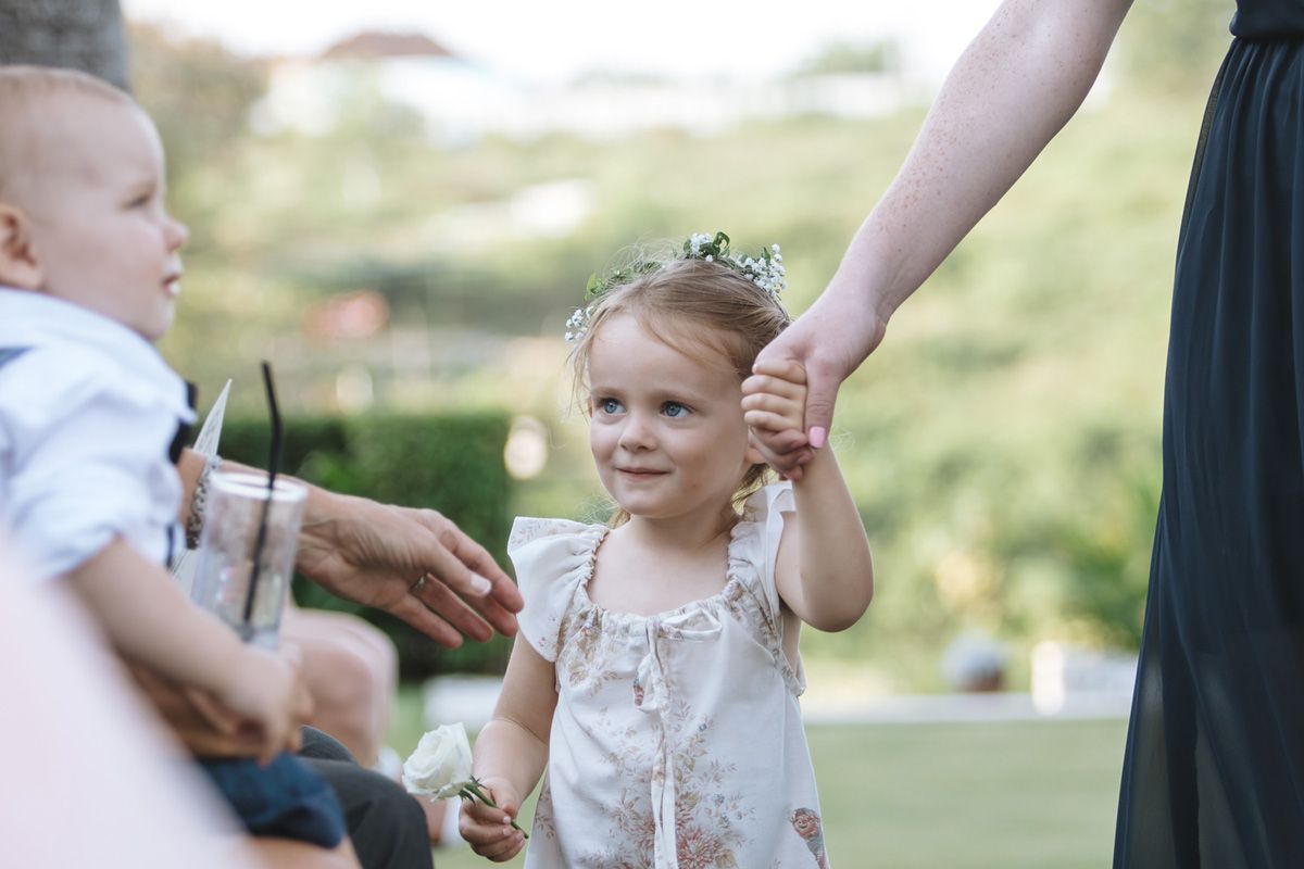 Small child in a white dress holds hands with an adult while walking outdoors in a sunny garden setting.