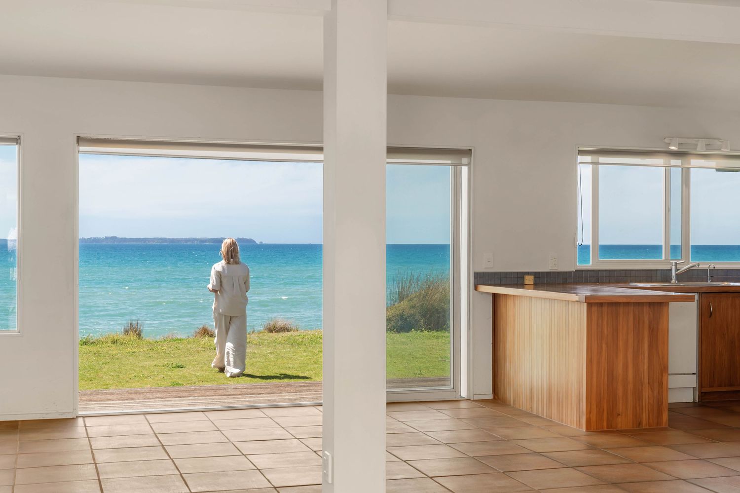 Modern beachfront interior with tiled floor and wooden counter offering panoramic ocean views through large windows.