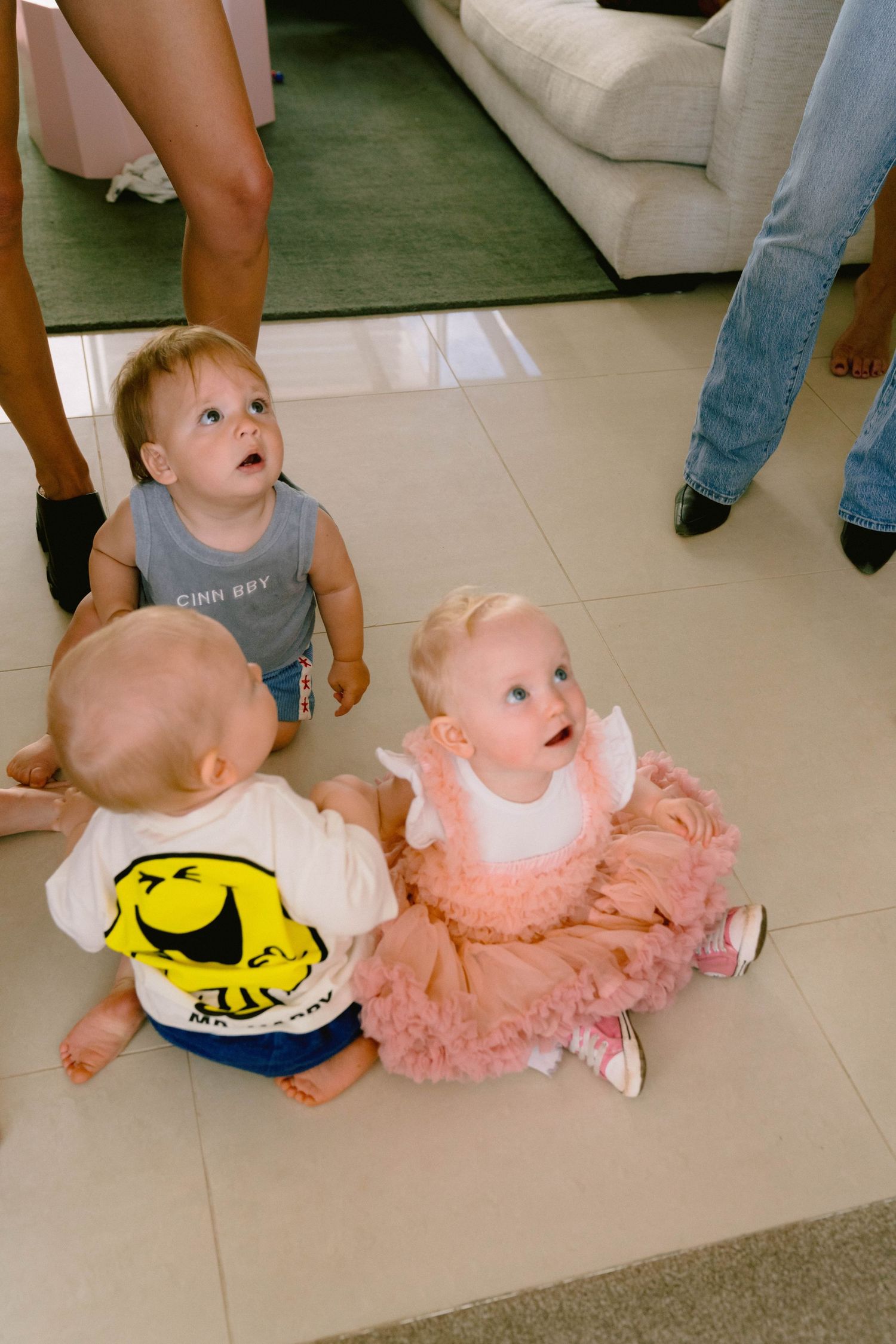 Two babies in coordinating outfits sitting on a white tiled floor during a party.