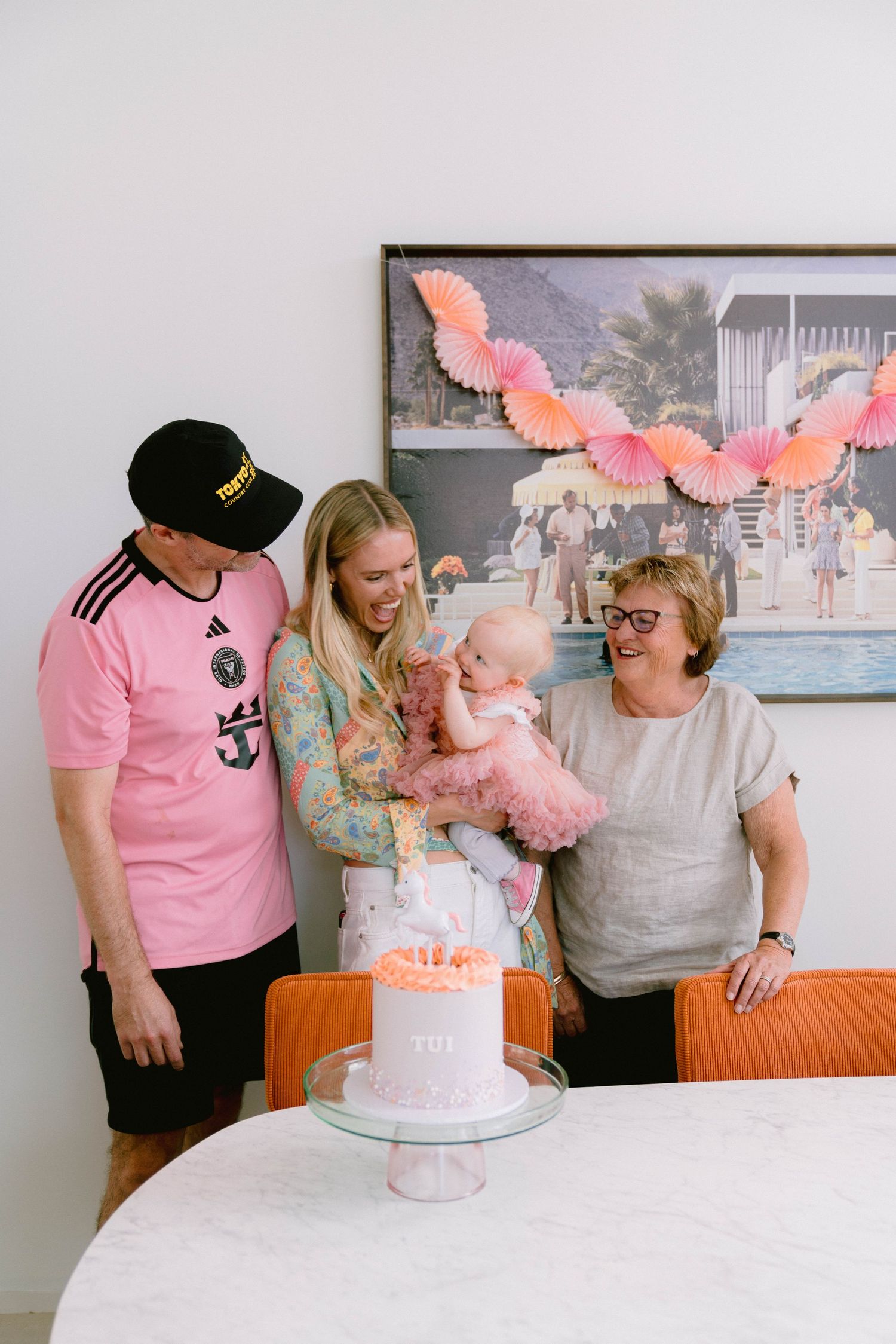 A birthday party celebration with a pink cake on a table and pink paper decorations hanging from above.