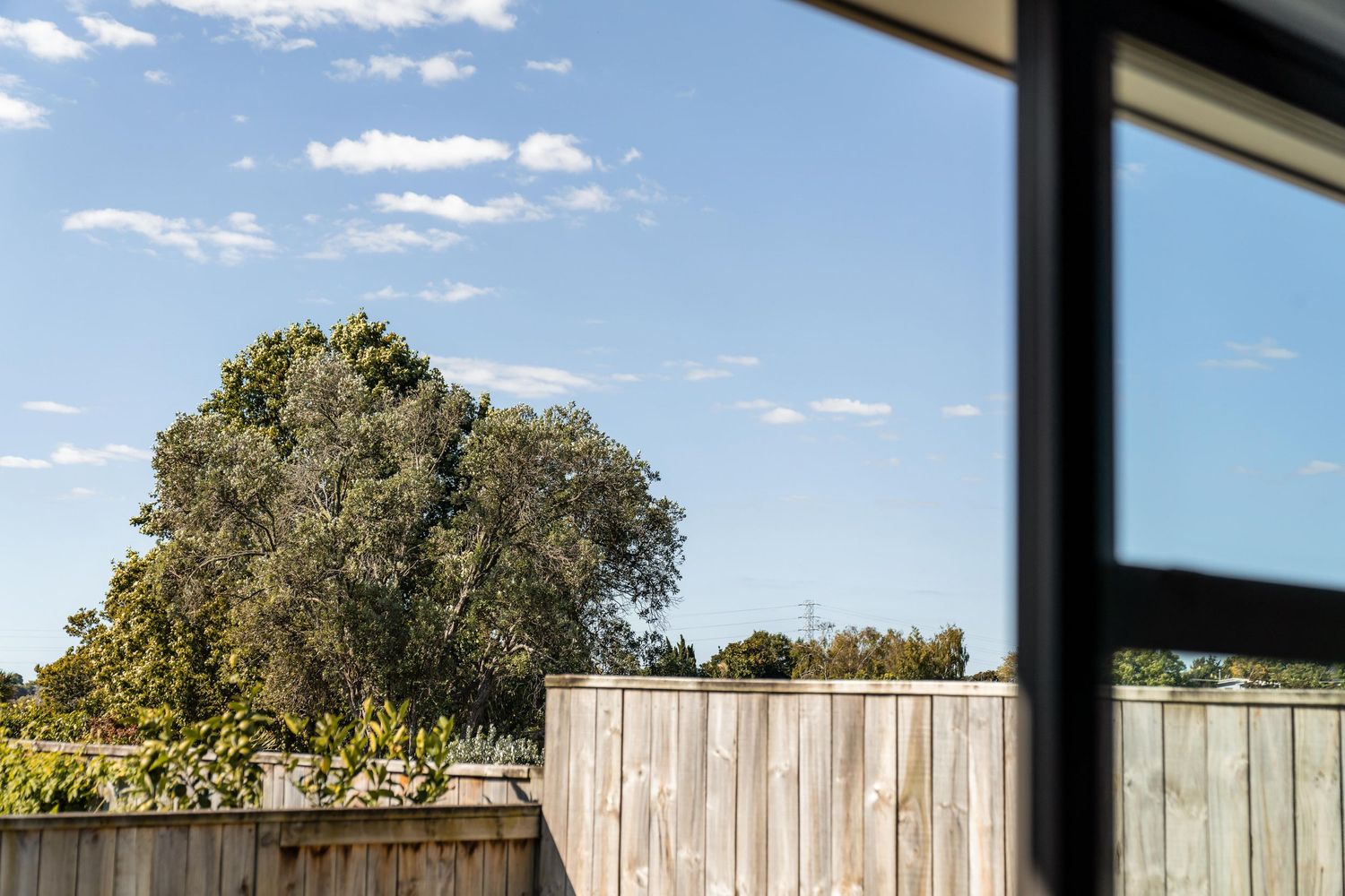 Scenic view through black-framed windows showing blue sky, trees, and wooden fence in contemporary bedroom setting.