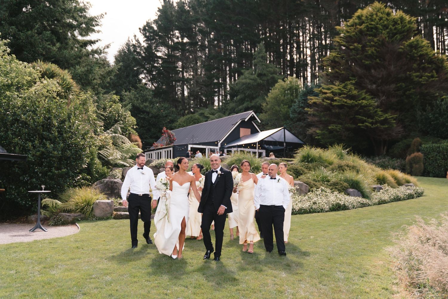 A wedding party walks together on a lush green lawn surrounded by tall pine trees and gardens.