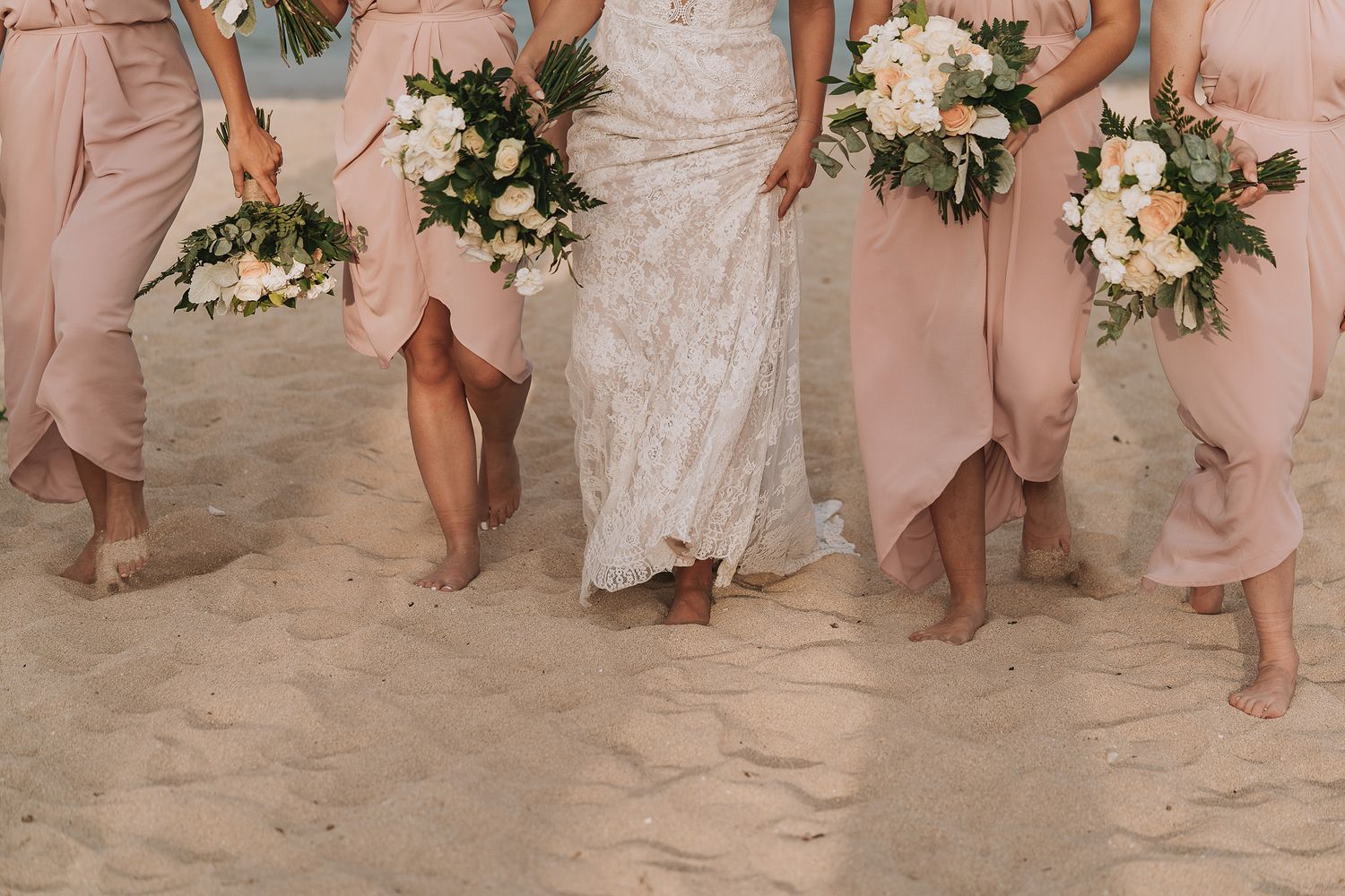 Bridesmaids in blush pink dresses holding floral bouquets stand barefoot on sandy beach.