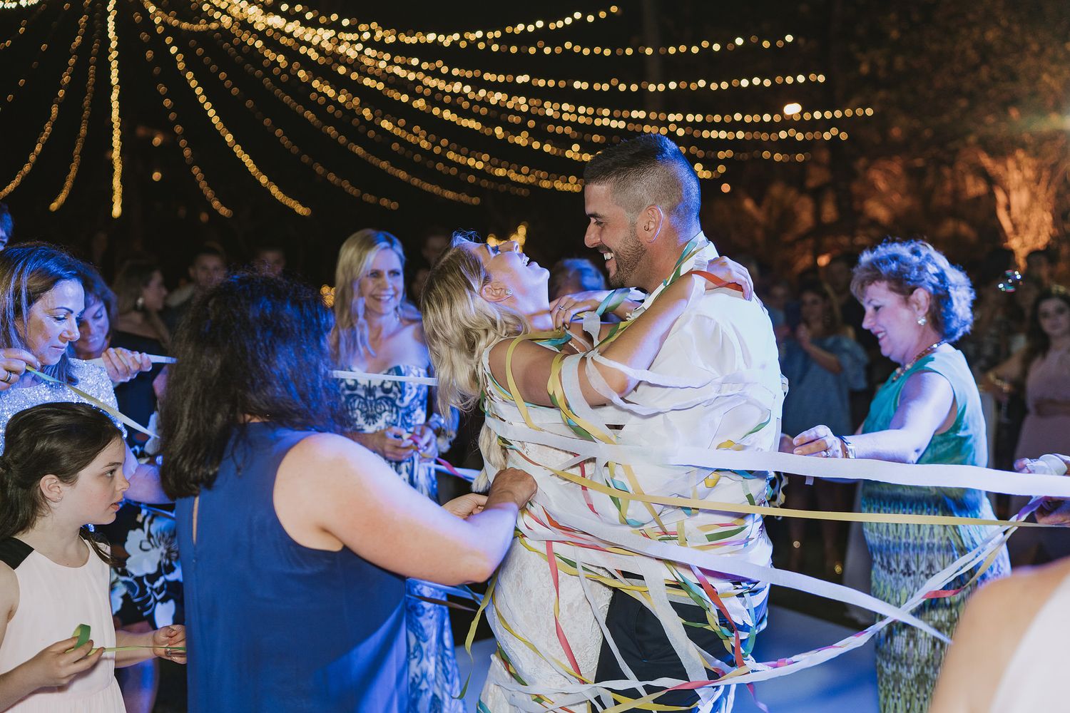 Wedding guests enjoying dancing and celebrations under twinkling string lights at an outdoor evening reception.