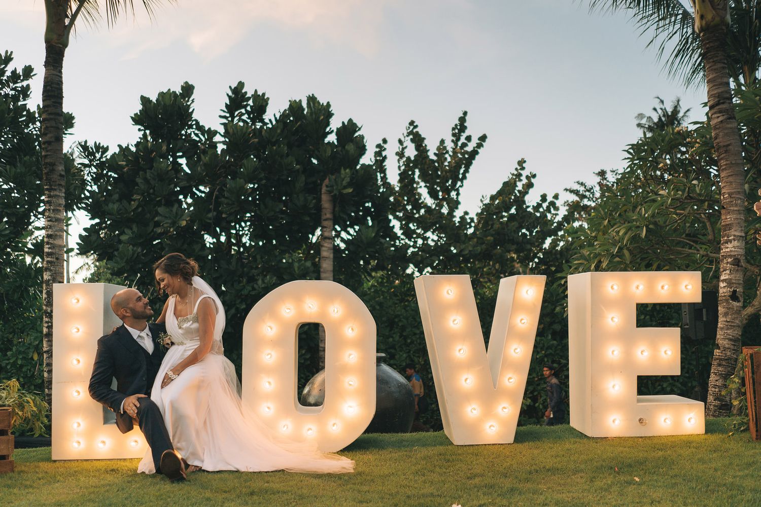 Large illuminated LOVE letters create a romantic backdrop against a natural outdoor setting at dusk with palm trees and greenery.