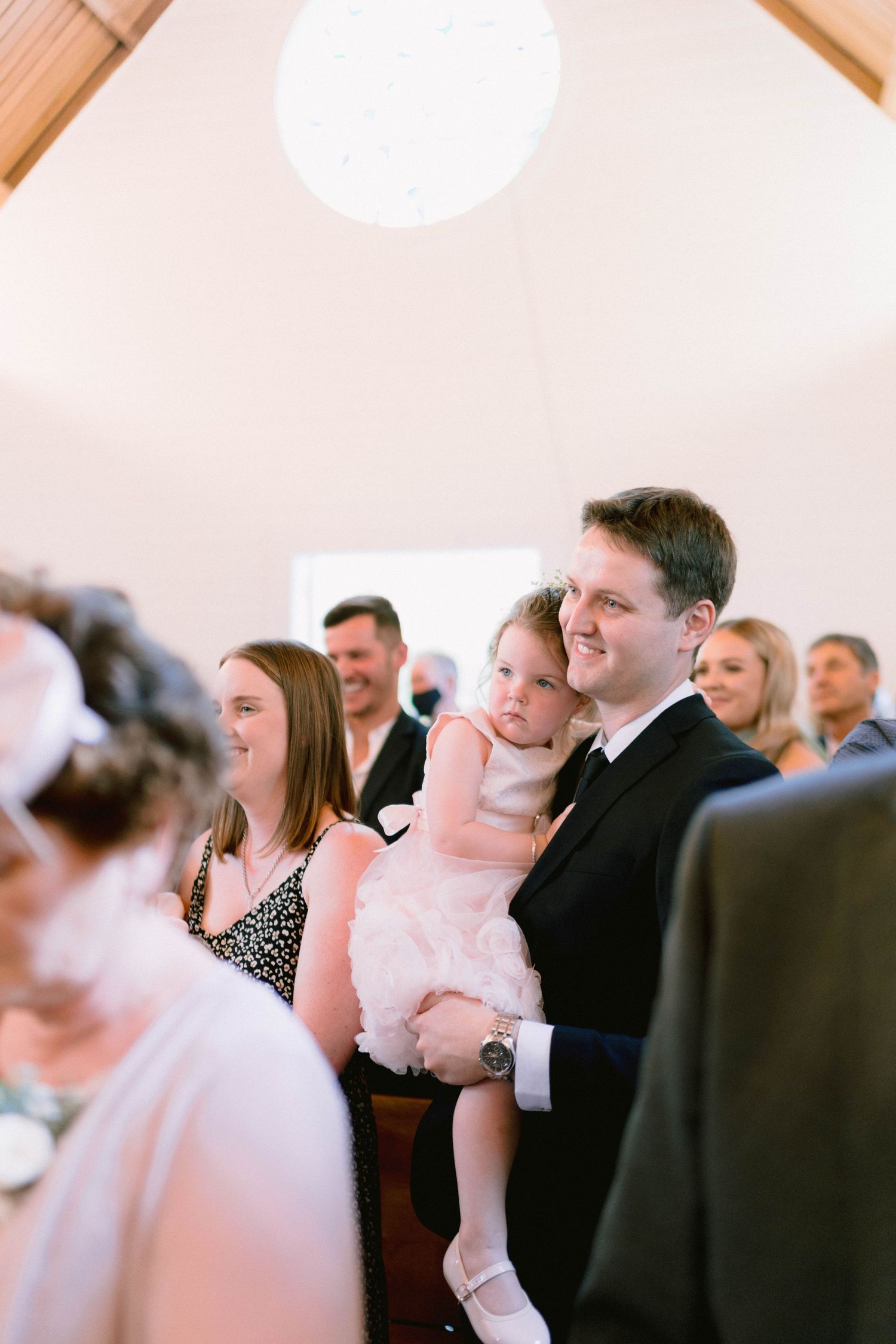 Wedding guests seated in church pews during ceremony with soft lighting from above.