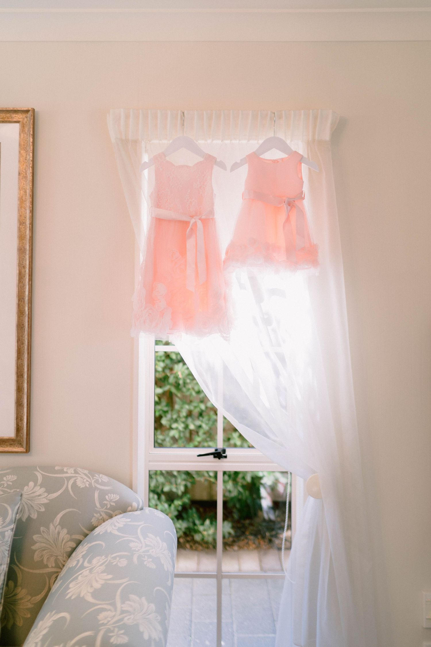 Two peach-colored dresses hang in front of a sunny window with white sheer curtains beside an upholstered chair.