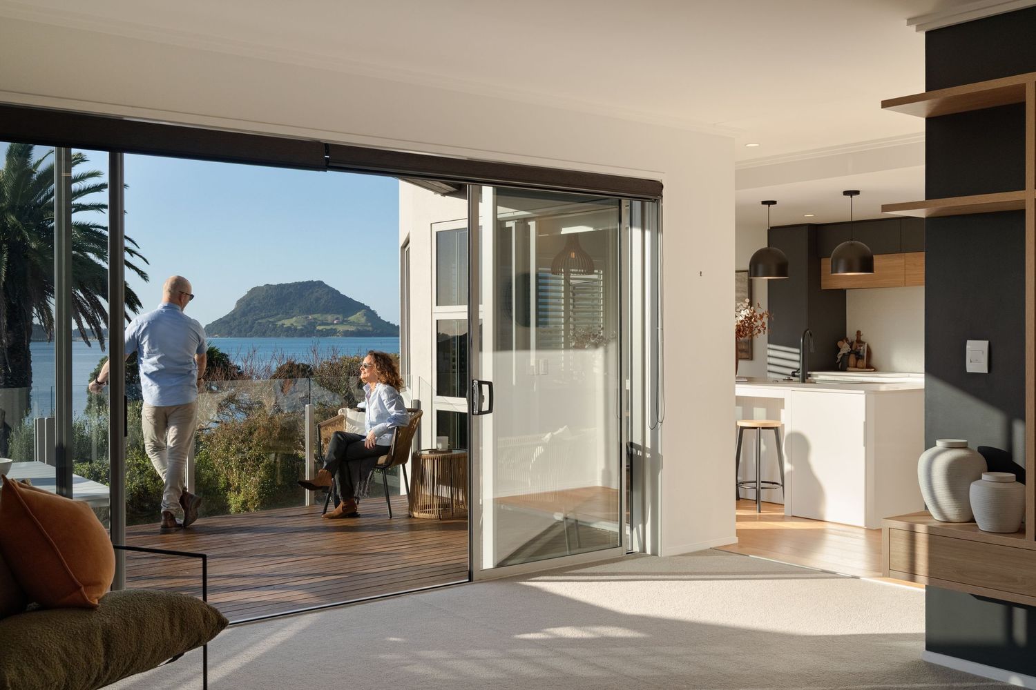 Modern open-plan living room with bifold doors opening to a deck with ocean views and two people relaxing outside.