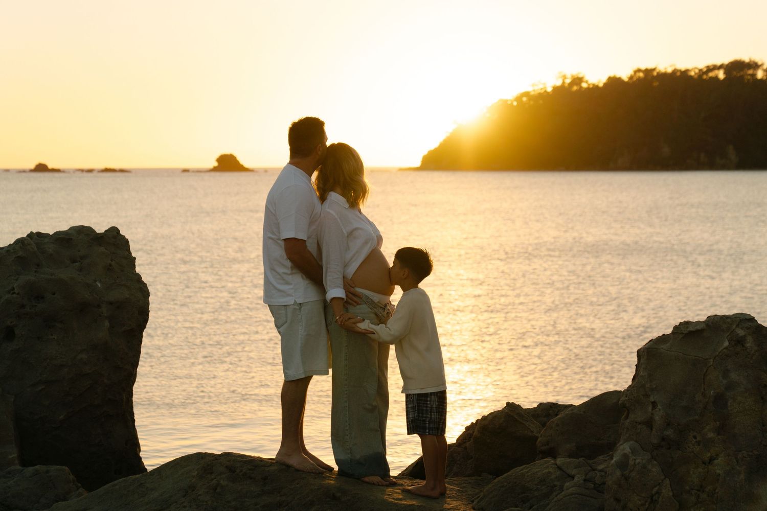 Family of three silhouetted against a golden sunset on rocky beach, watching the ocean horizon together. Bryony Alexander Newfound.