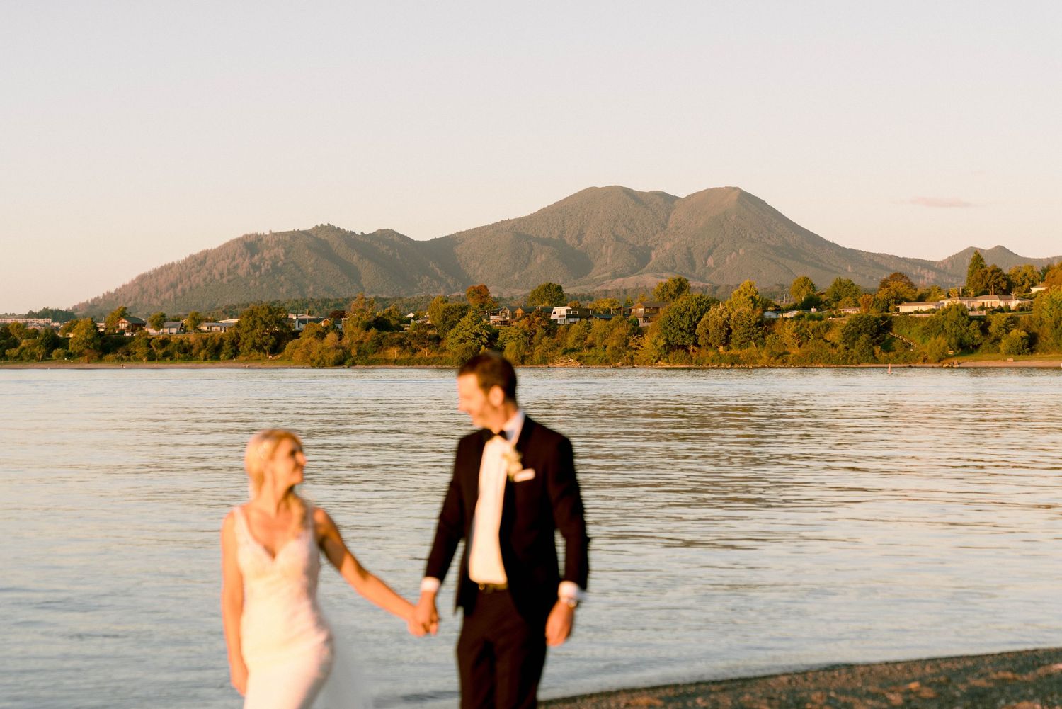 A couple walks along a scenic lakeside beach at sunset with mountains in the background.