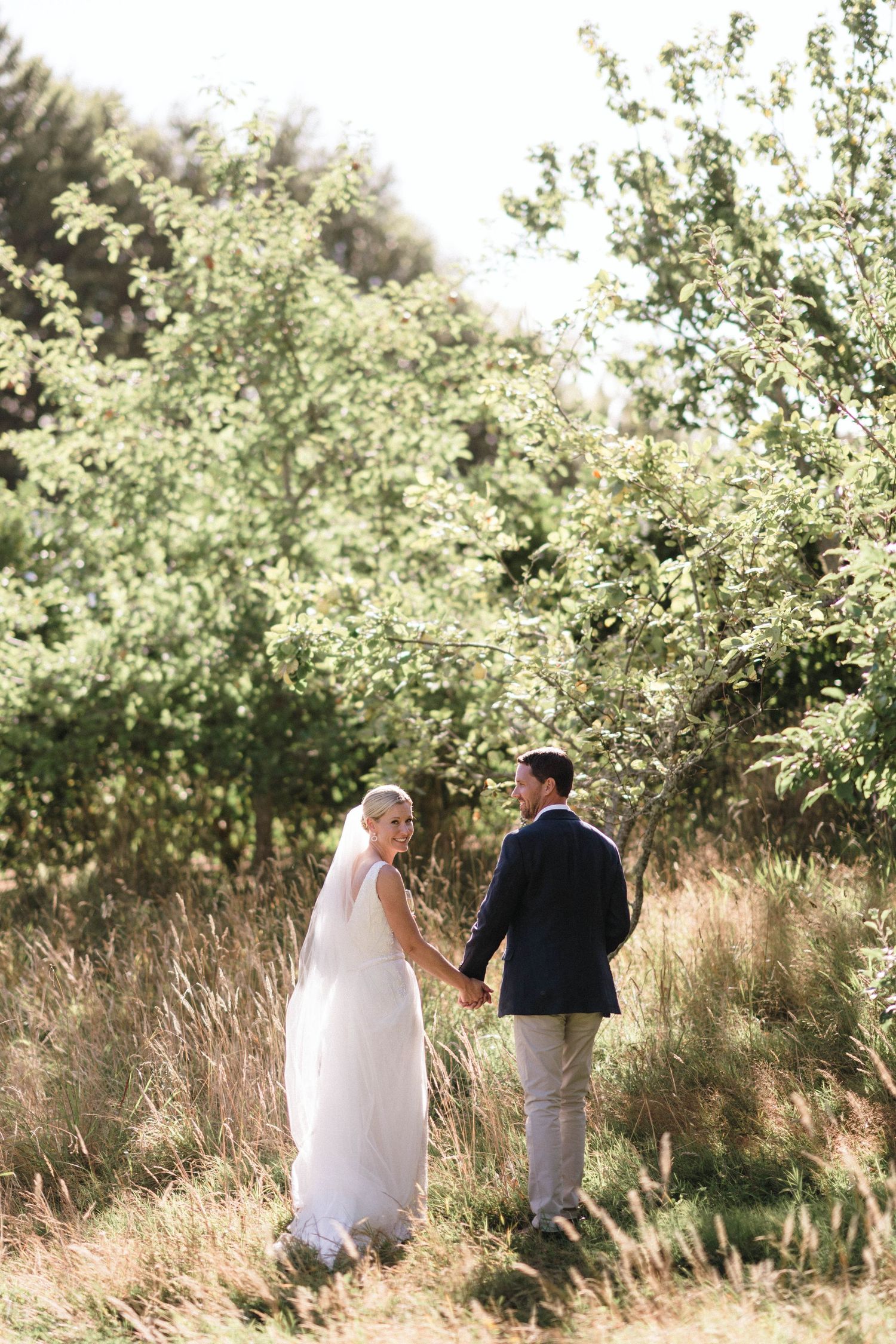 A couple walks hand in hand through tall grass and flowering trees on their wedding day.
