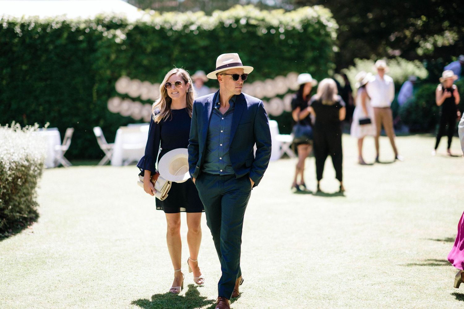 A stylish couple strolls together at an outdoor social event with other attendees visible in the background.