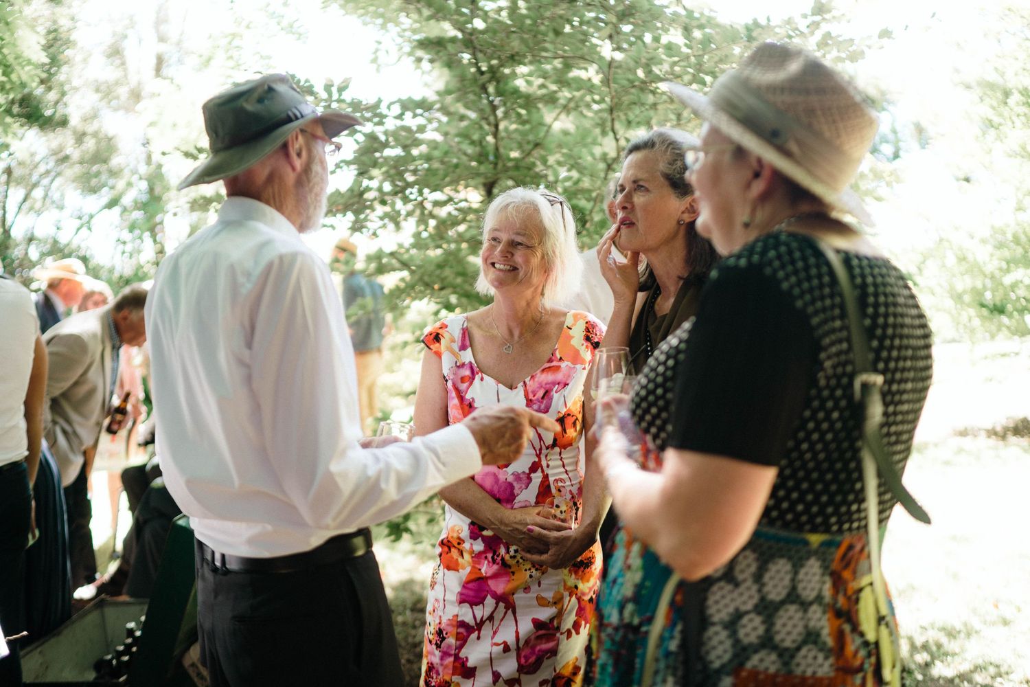 A group of people chatting and laughing together at an outdoor social gathering in a sunny wooded setting.