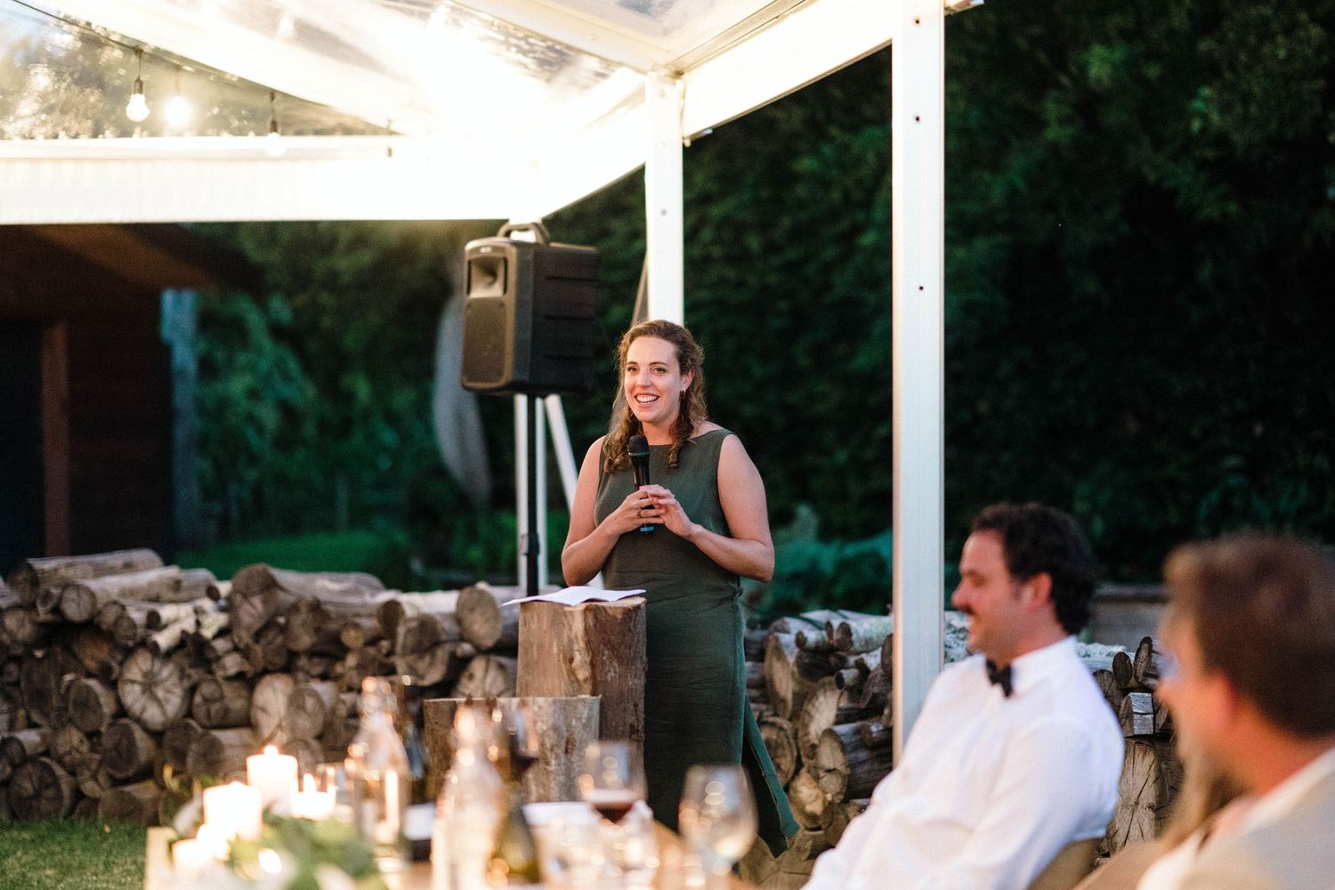A person gives a speech under a white outdoor pergola during an evening wedding reception dinner.