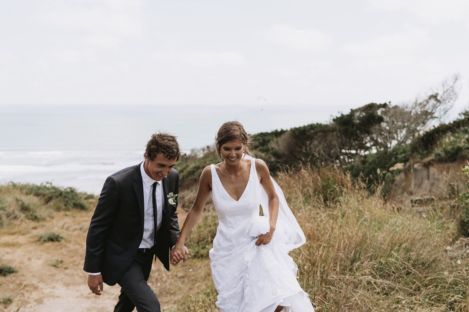 A couple walks together through tall grass along a scenic coastal path on their wedding day.