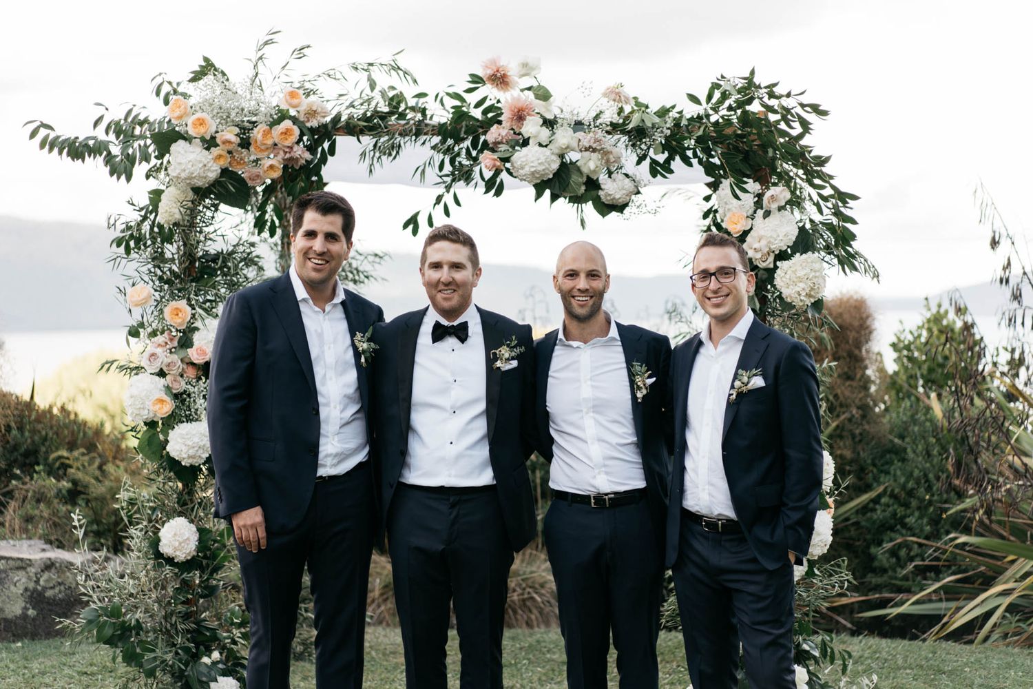 Groomsmen in black tuxedos and bowties laughing together outdoors at sunset.