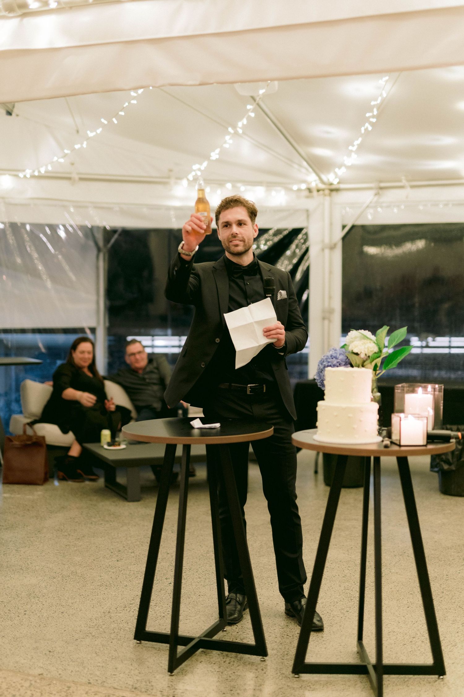 A speaker in black attire gives a toast at a wedding reception near decorative tables.