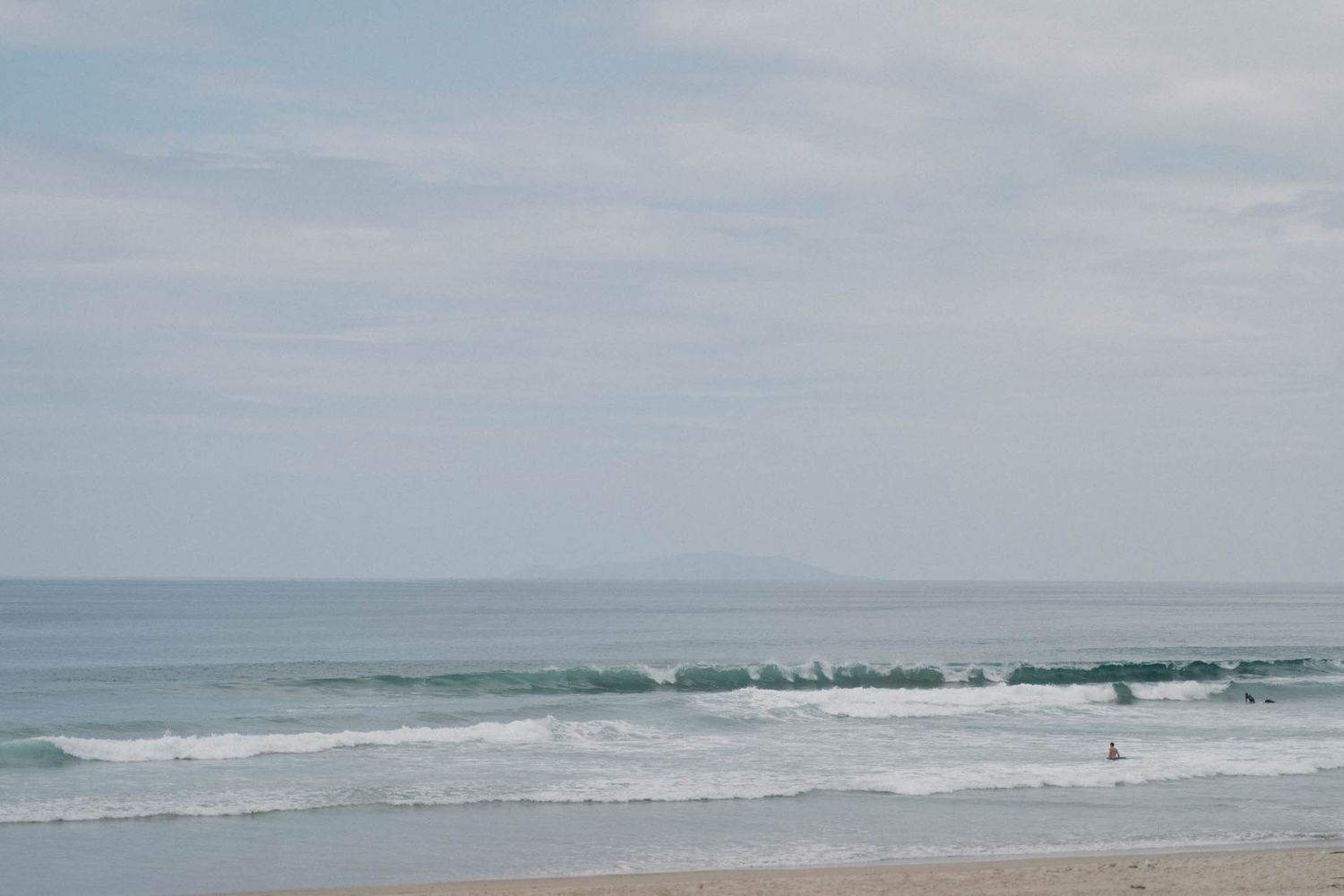 Sand dunes stretch along a cloudy beach coastline with waves rolling onto the shore at the water's edge.