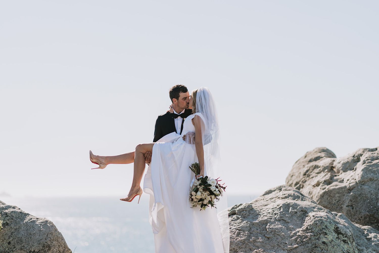 A romantic coastal wedding photo with a couple embracing on rocky cliffs by the ocean.