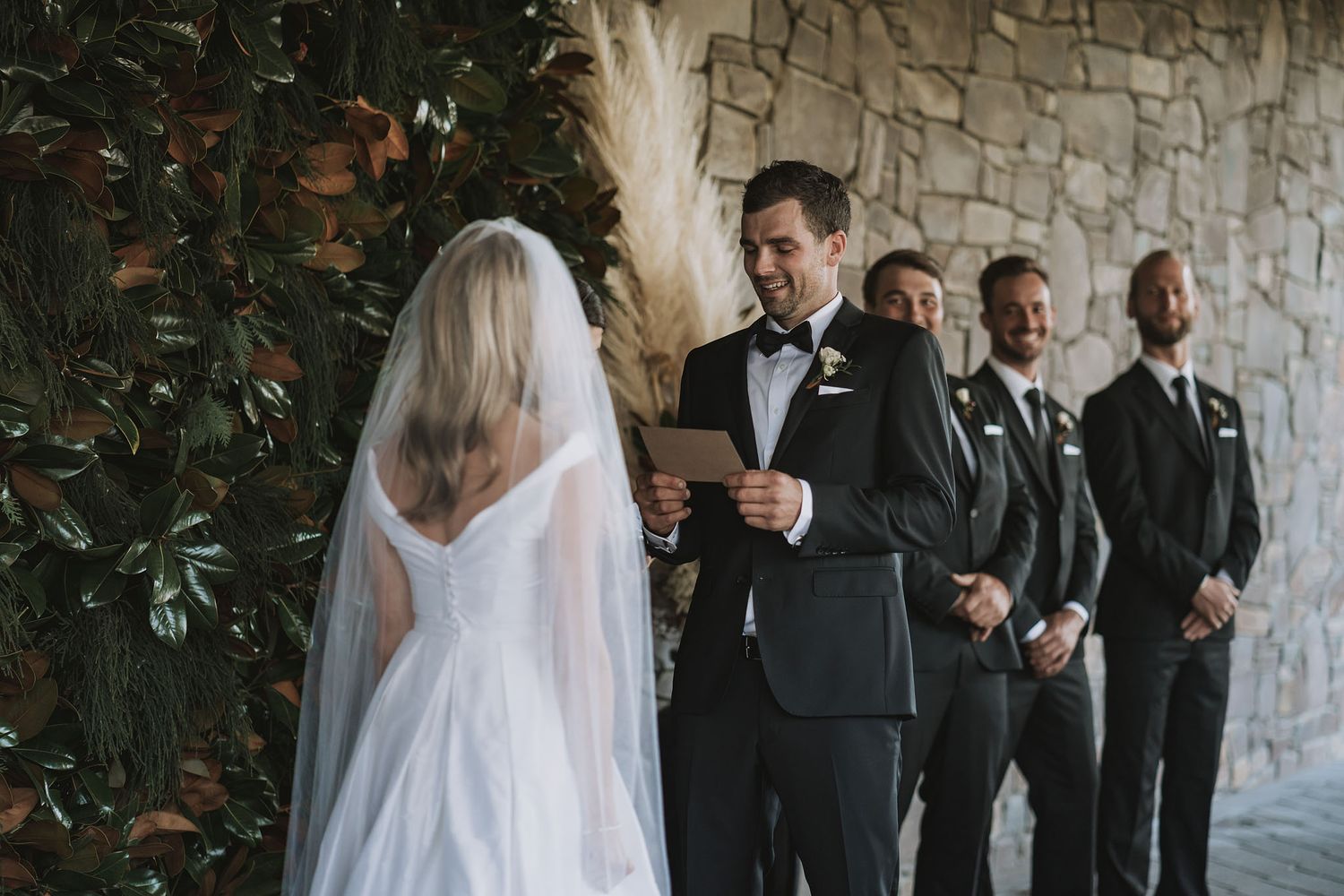 A couple exchanges vows during an outdoor wedding ceremony with groomsmen standing in dark suits against a stone wall.