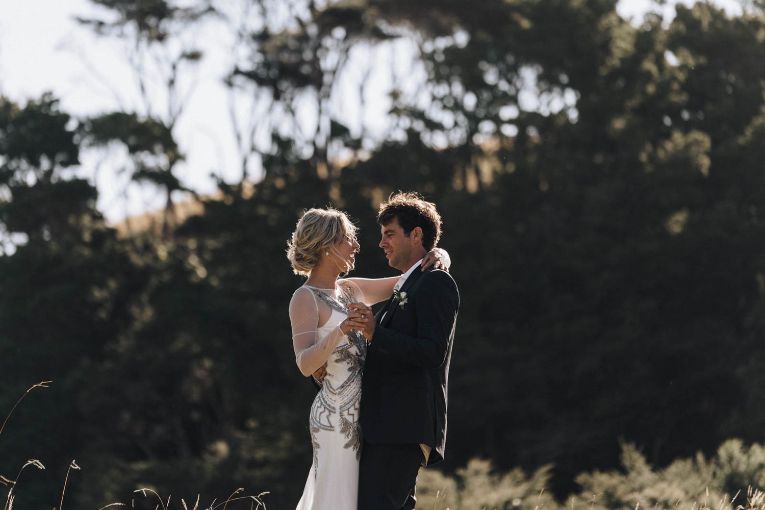 A couple in wedding attire shares an intimate moment at sunset with trees silhouetted in the background.