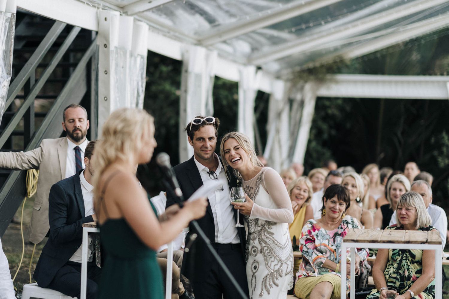 Wedding guests enjoying cocktail hour in outdoor tent with glass ceiling and white drapes.