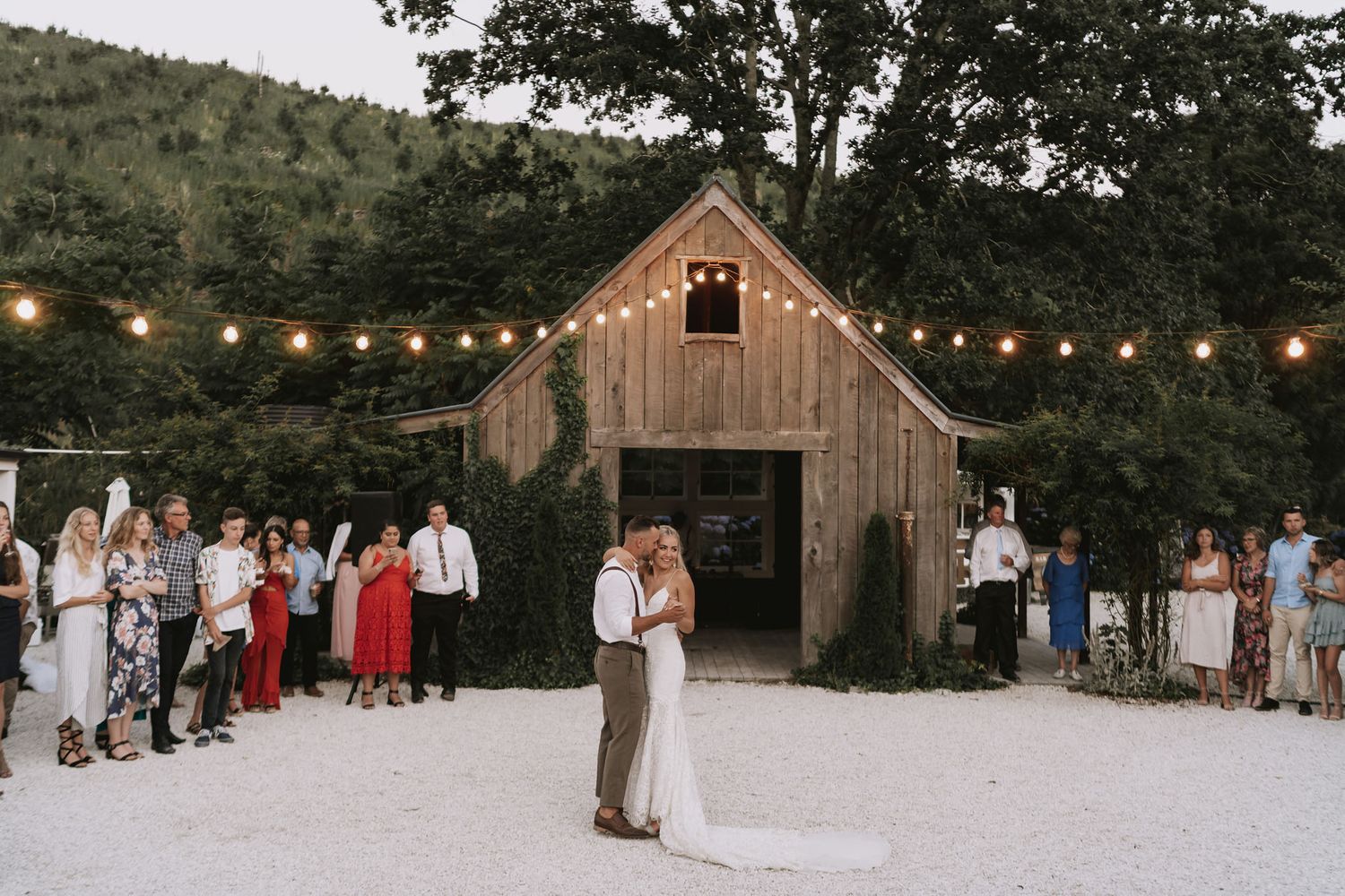 A couple shares their first dance on a rustic barn dance floor with string lights and guests watching.