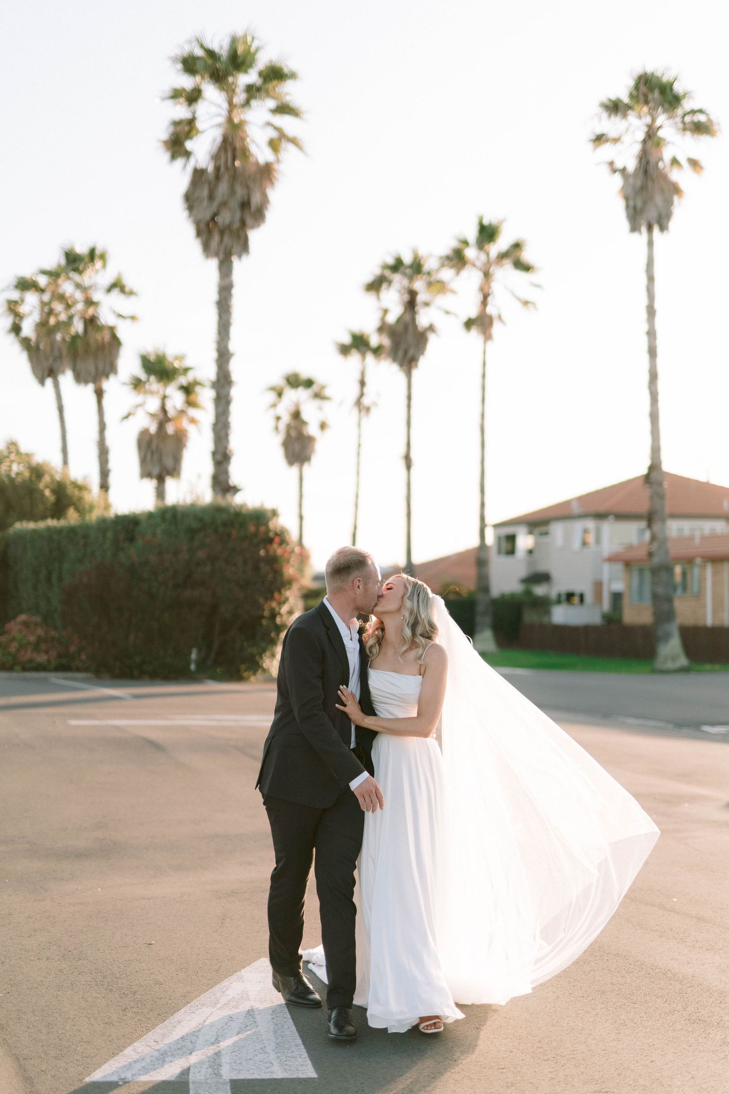 Newlyweds embrace during sunset with palm trees and Spanish-style buildings in the background.