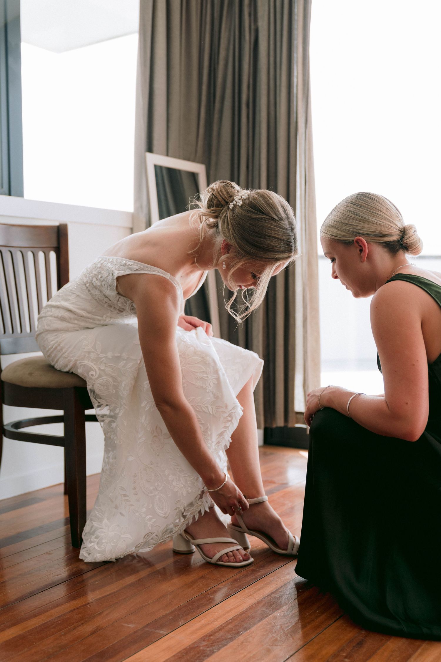 Bride in white dress putting on her wedding shoes while sitting on wooden floor.