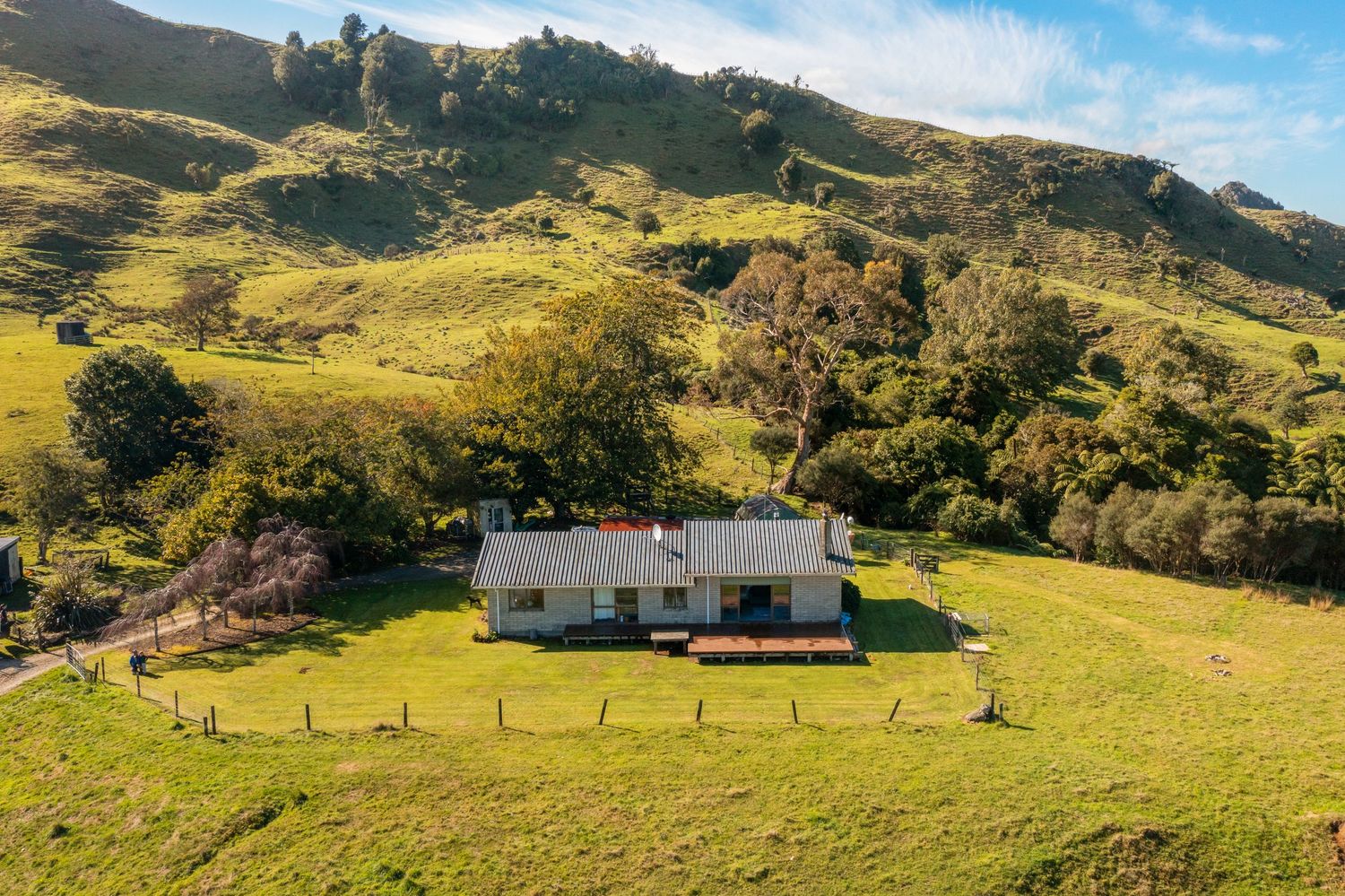 White farmhouse nestled in rolling green hills with mountain backdrop and wooden fencing.