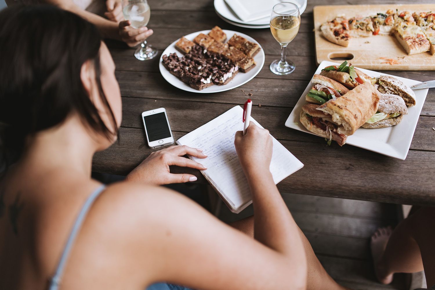 Food spread on wooden table with phone and notebook, featuring appetizers and wine glasses.
