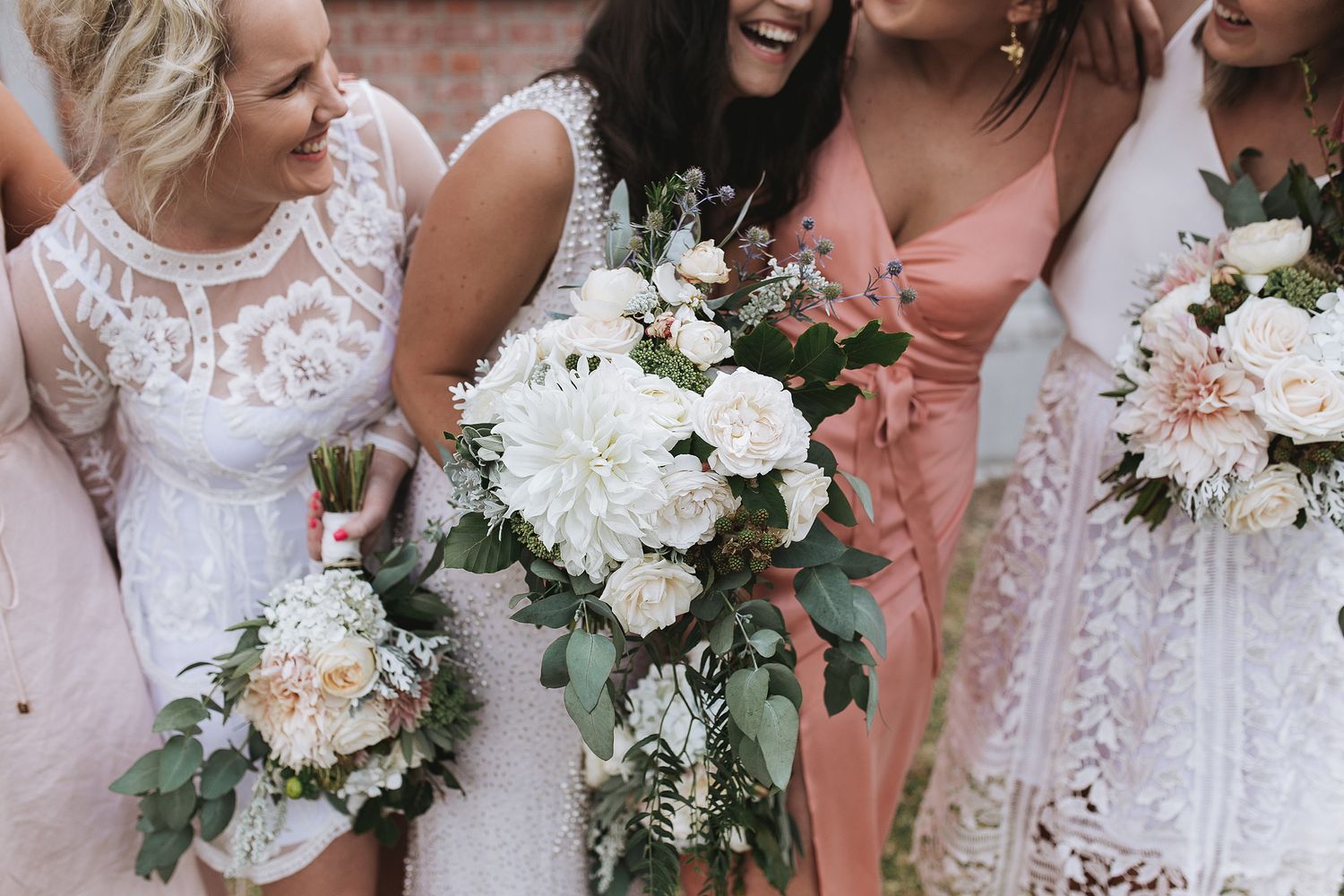 Bridesmaids in white lace and pink dresses hold elegant white and blush floral bouquets with greenery.