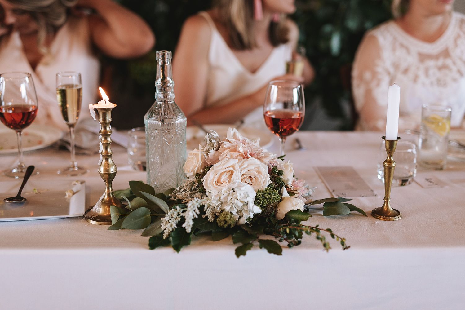 Elegant wedding table setting with pink floral centerpiece, brass candlesticks, and glassware on white tablecloth.