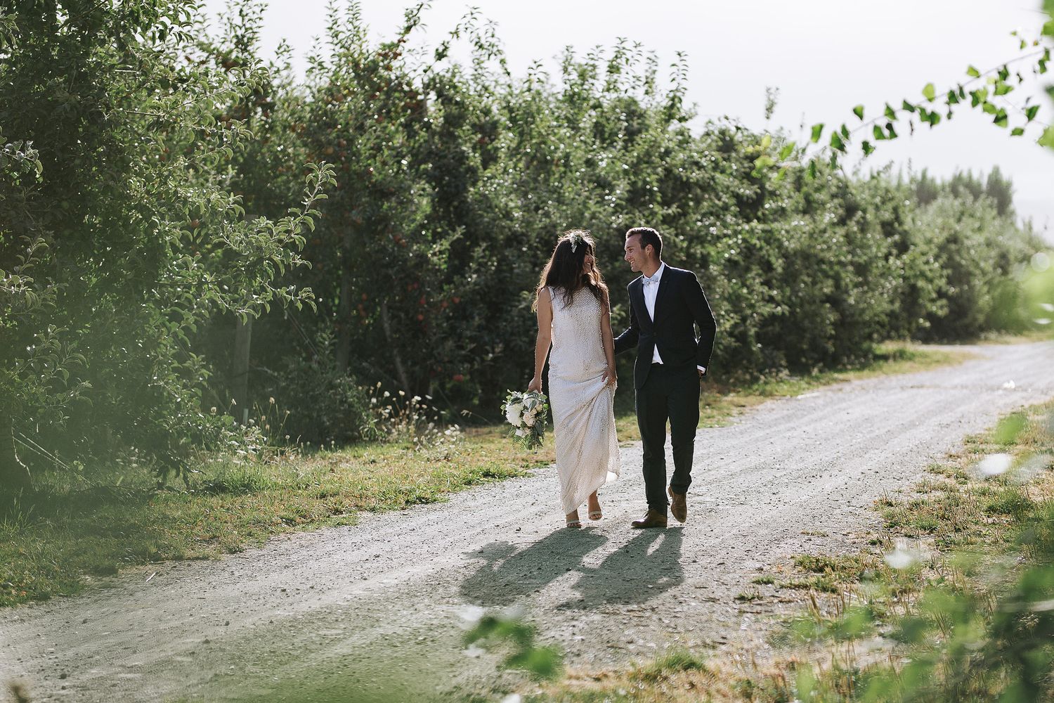 Two figures walk together on a sunny gravel path lined with lush green foliage and trees.