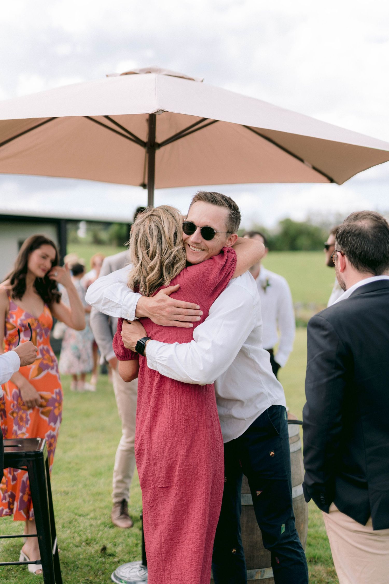 Wedding guests sharing warm embraces during an outdoor reception under beige umbrellas.